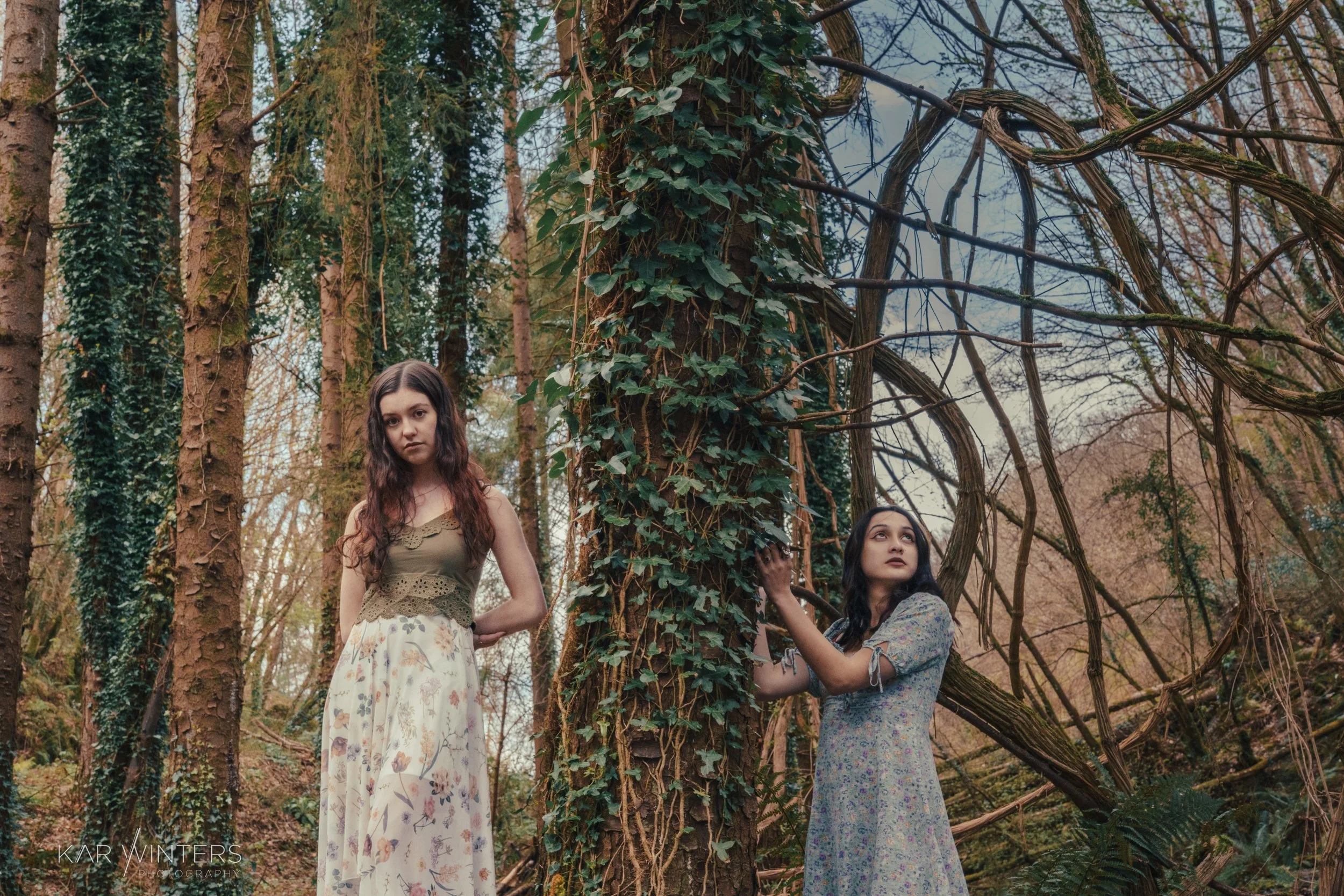 Two young women in long dresses in a forest with tall trees and vines, one touching a large tree trunk, both looking contemplative.