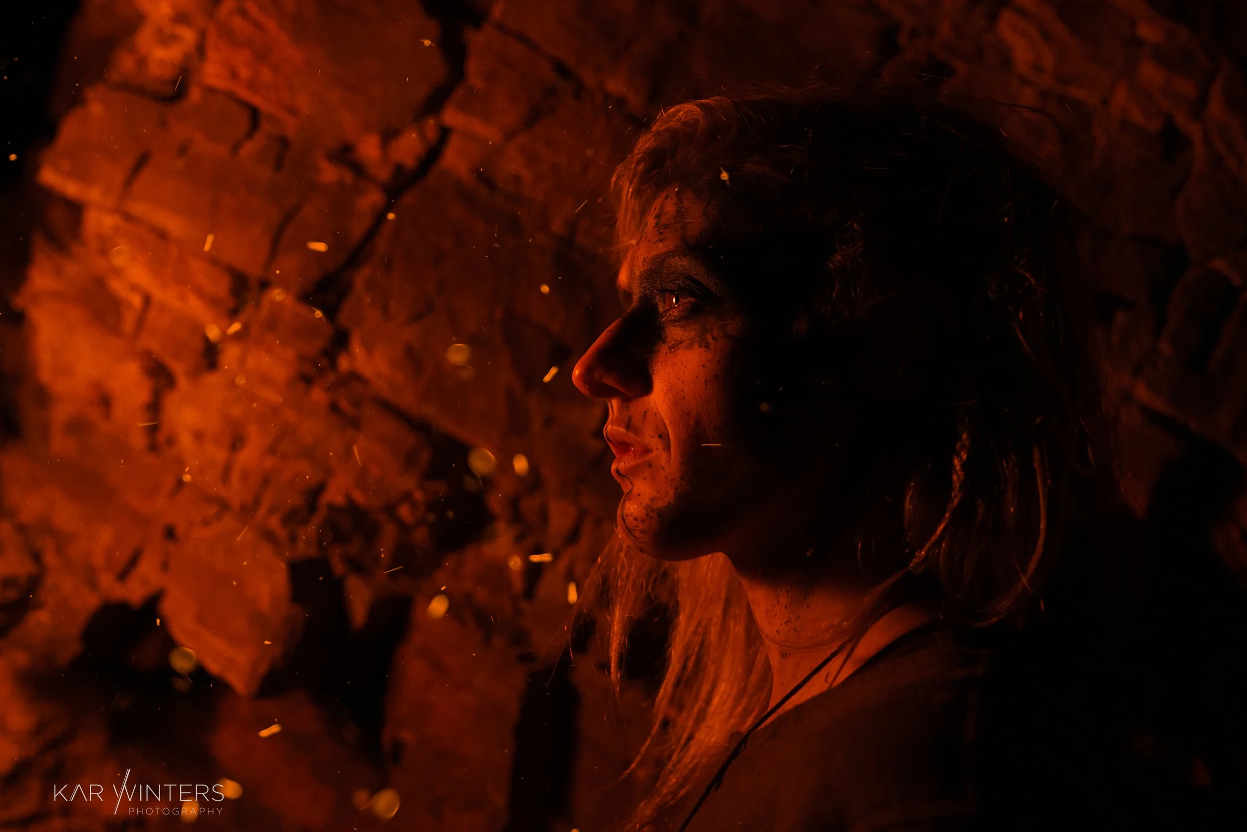 A woman with long hair and dirt on her face, illuminated by orange firelight, sitting against a brick wall, with visible sparks or embers floating around.