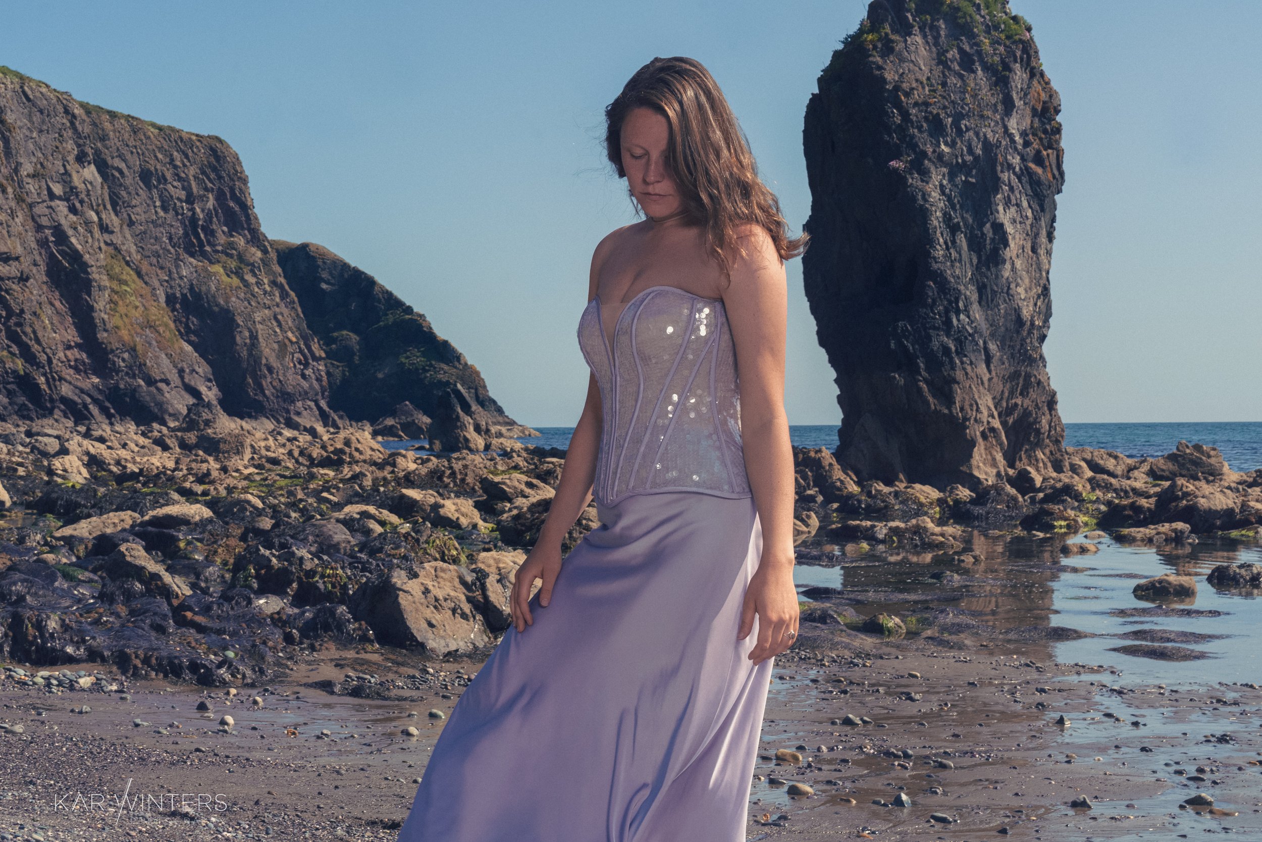 A woman in a pale lavender dress with a strapless, fitted bodice stands on a rocky beach with large rock formations in the background, under a clear blue sky.