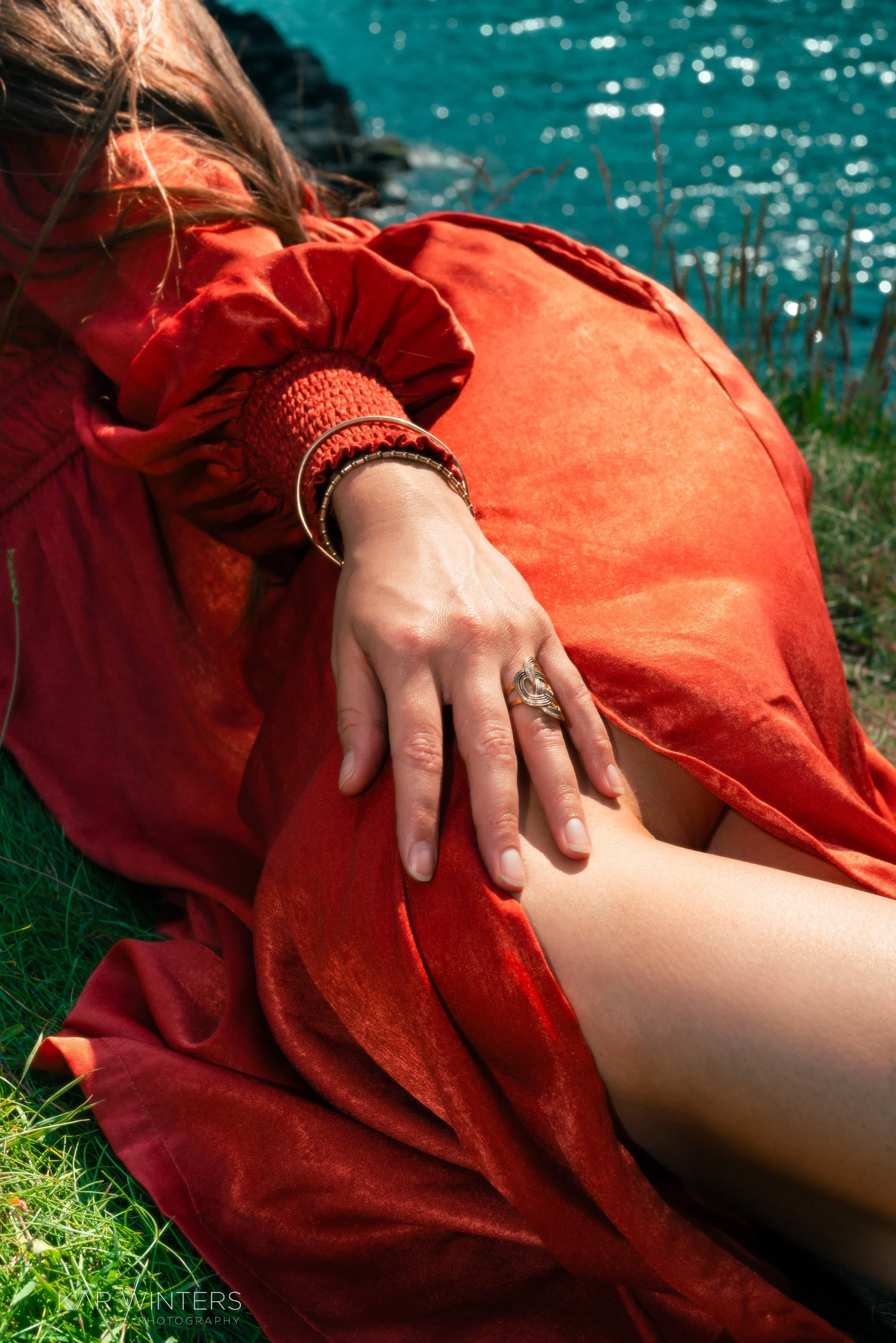 Close-up of a woman wearing a red dress with jewelry, sitting on grass near a body of water.