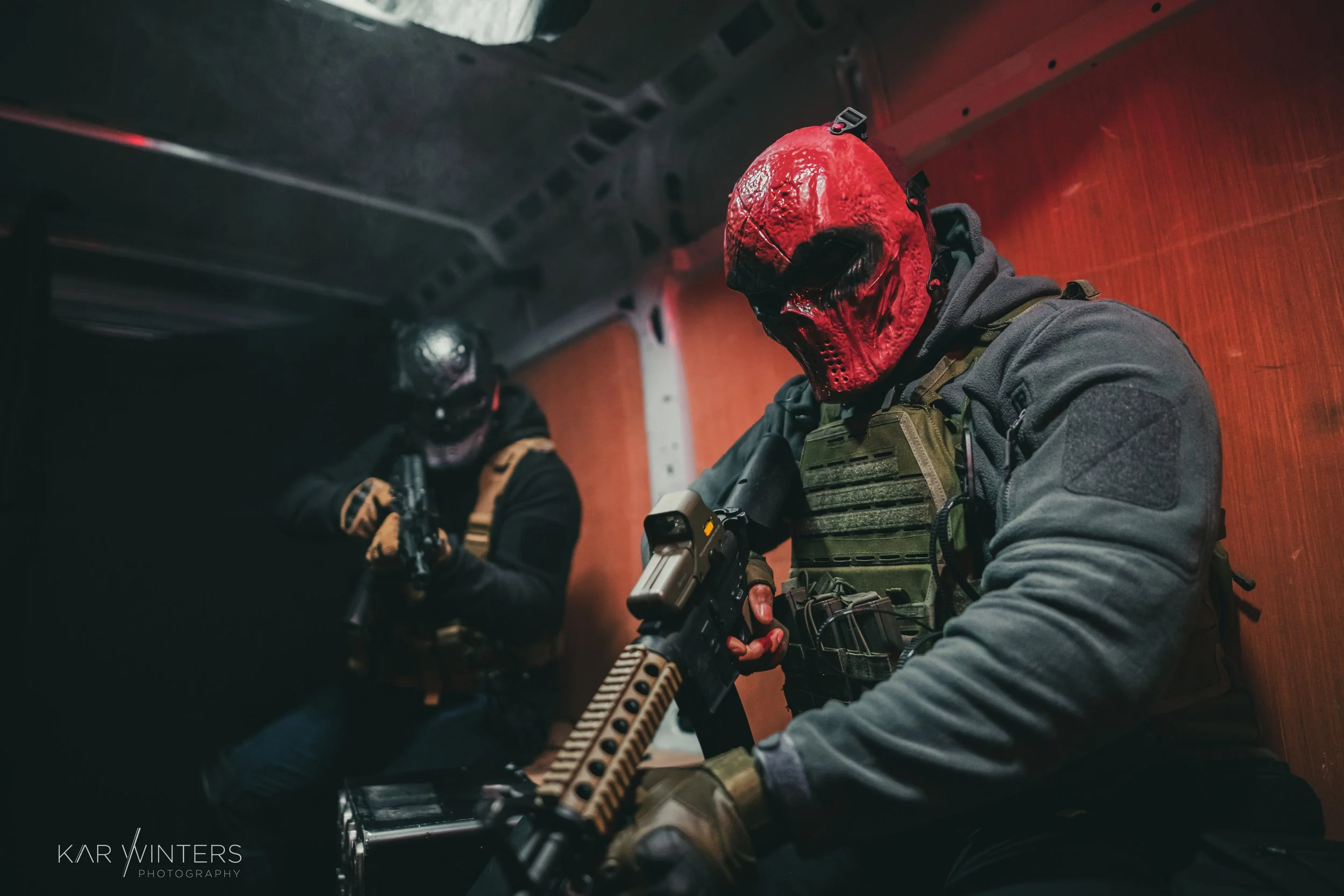 Two individuals wearing tactical gear and red skull masks inside a confined space, one holding a handgun and the other with a rifle, with a wooden wall background.