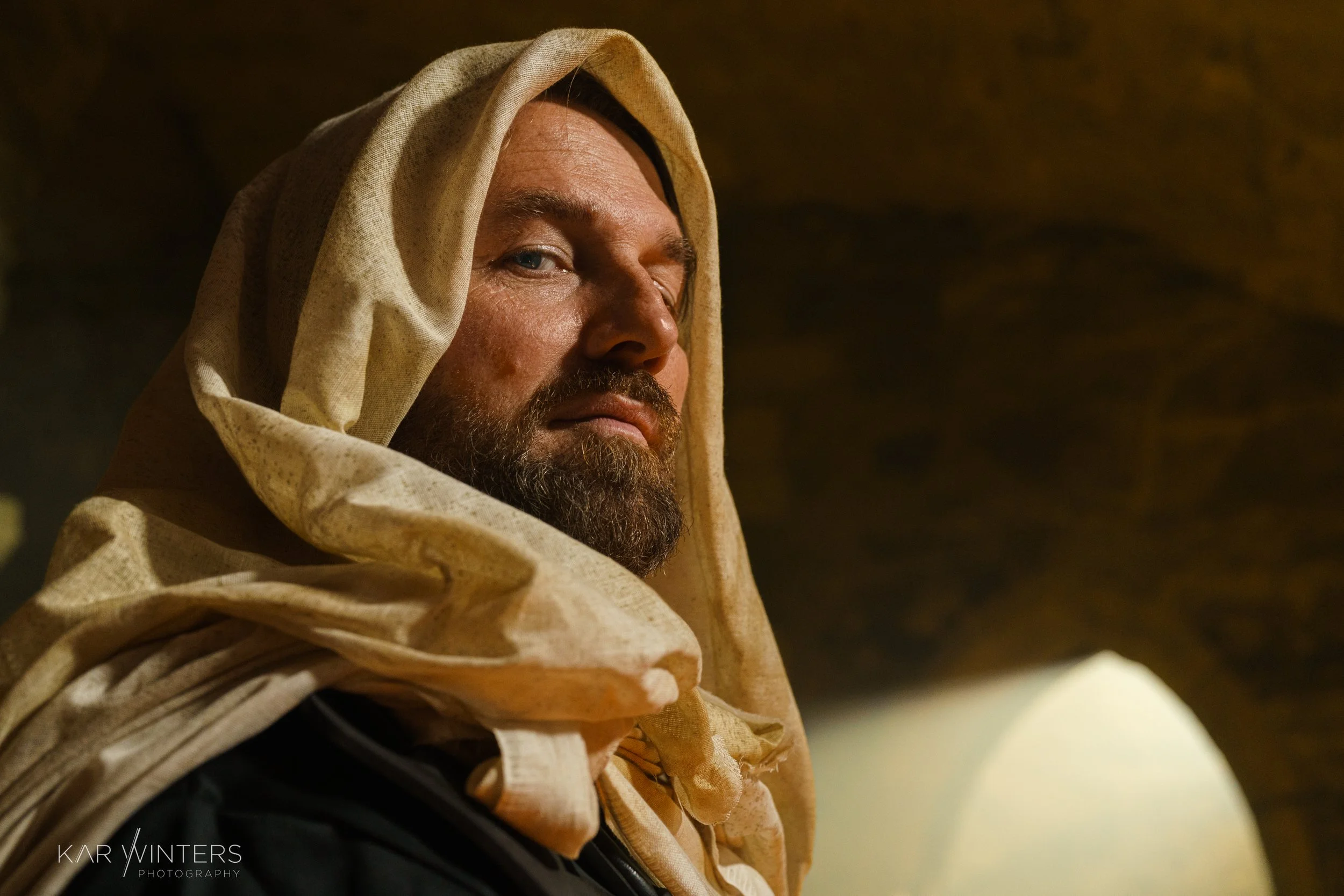 A man with a beard wearing a beige headscarf, looking over his shoulder in a setting with arched stone walls.