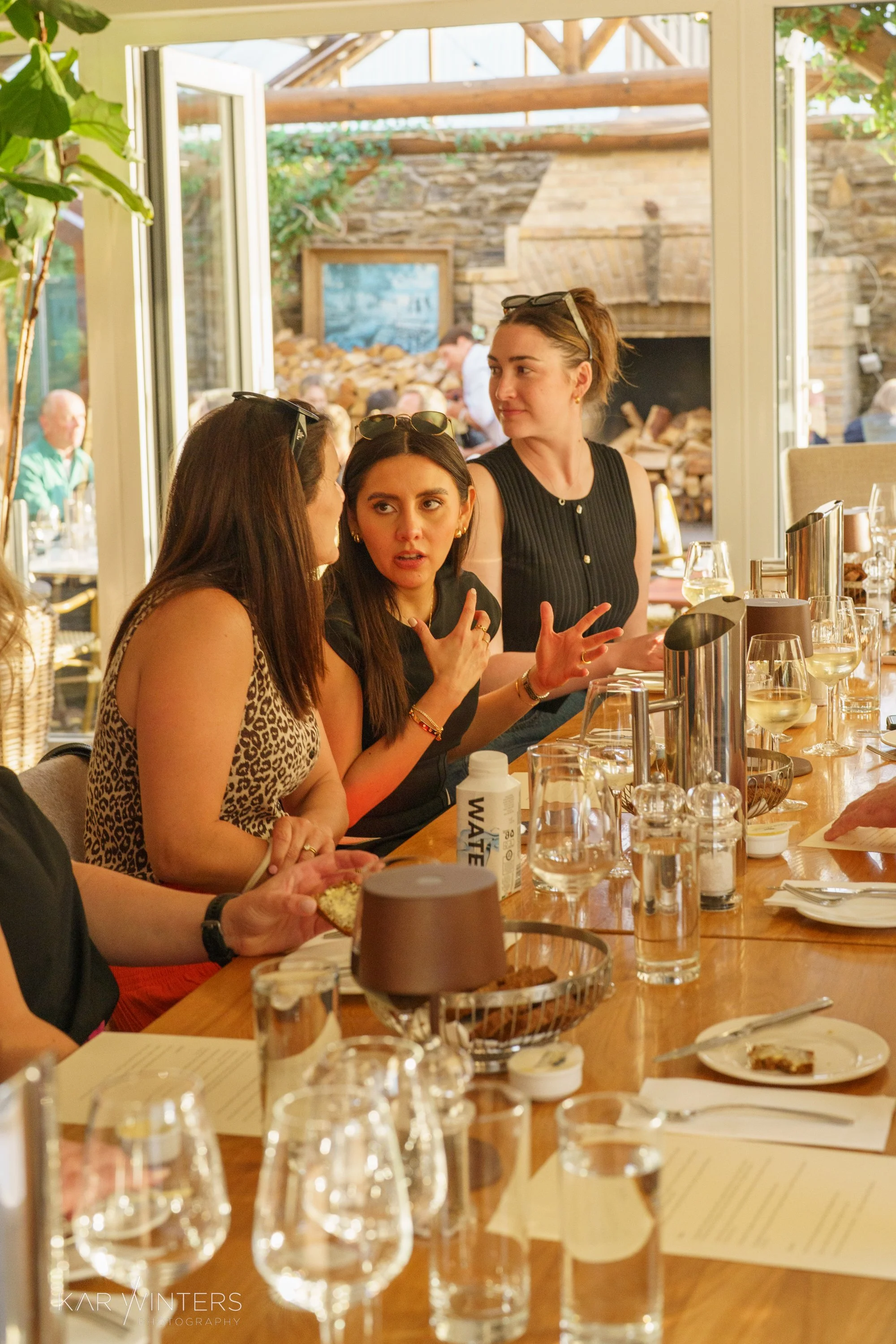 People having a conversation at a dining table in a restaurant, with glasses, plates, and bottles in front of them, and an open door leading outside to a patio or garden area.