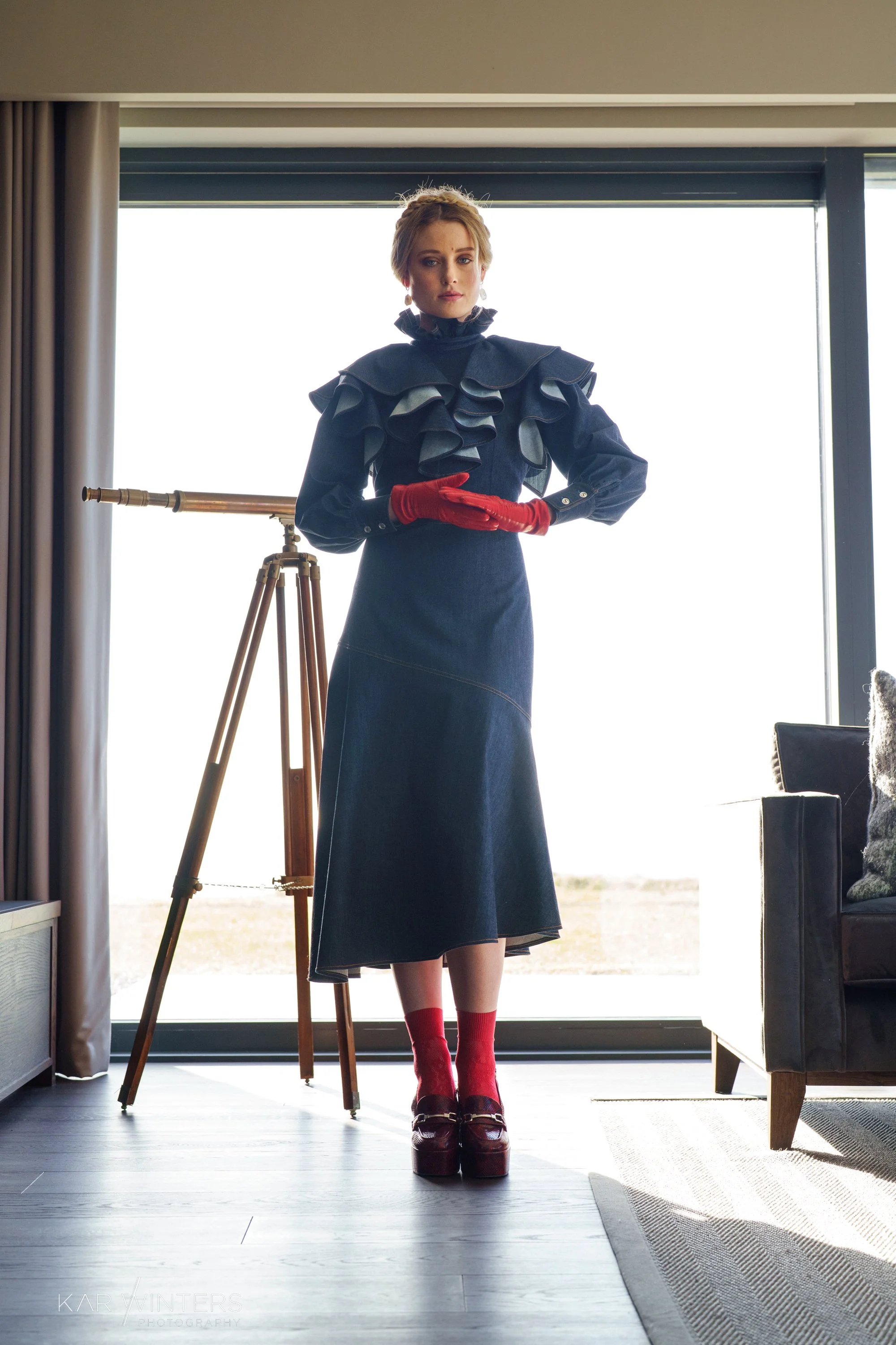 A woman stands inside a room in front of a large window, wearing a dark dress with ruffles on the shoulders, red gloves, red socks, and dark platform shoes. There is a telescope beside her and a couch to her right.