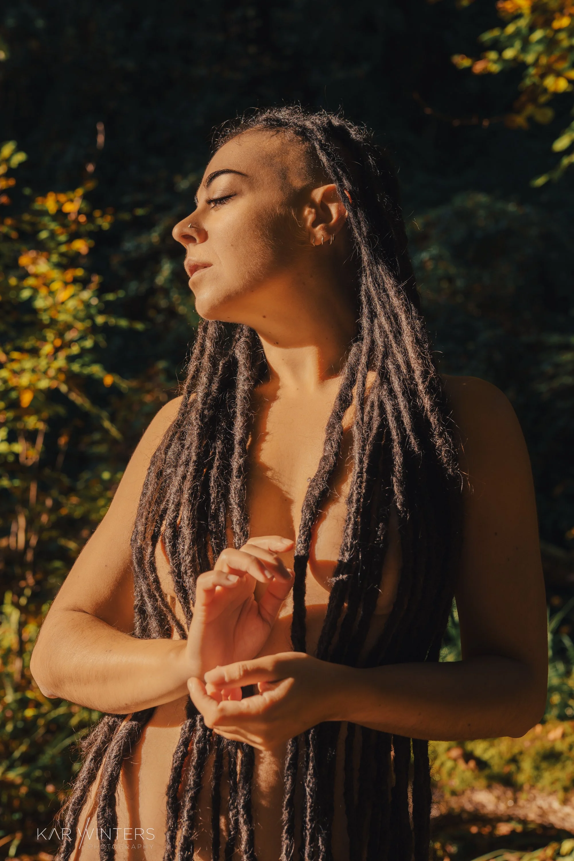 A woman with dreadlocks standing outdoors, eyes closed, appearing peaceful in sunlight, with a background of dark foliage.