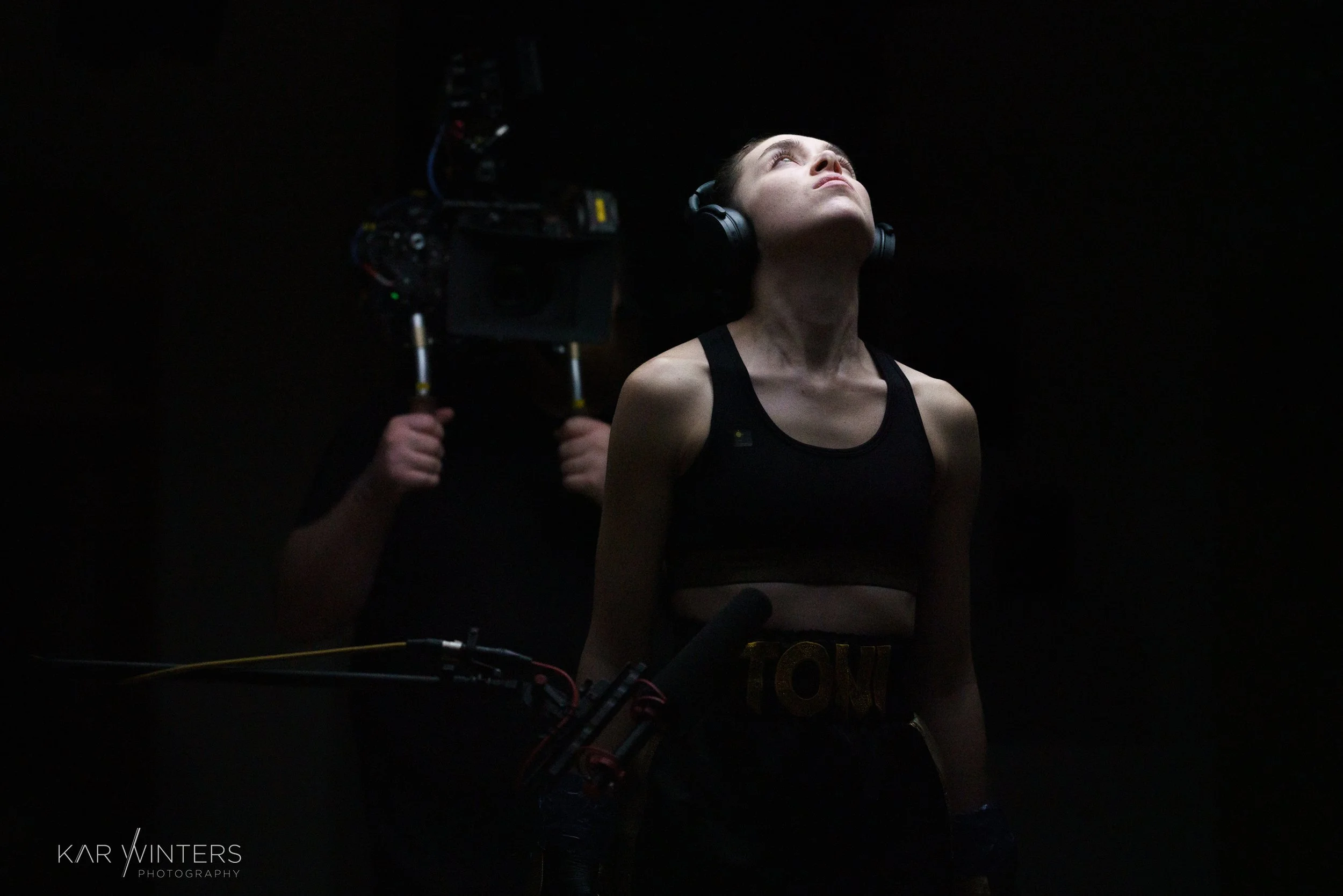 A woman wearing headphones and a sports bra looks upward in a dark environment, with a camera operator in the background recording her.