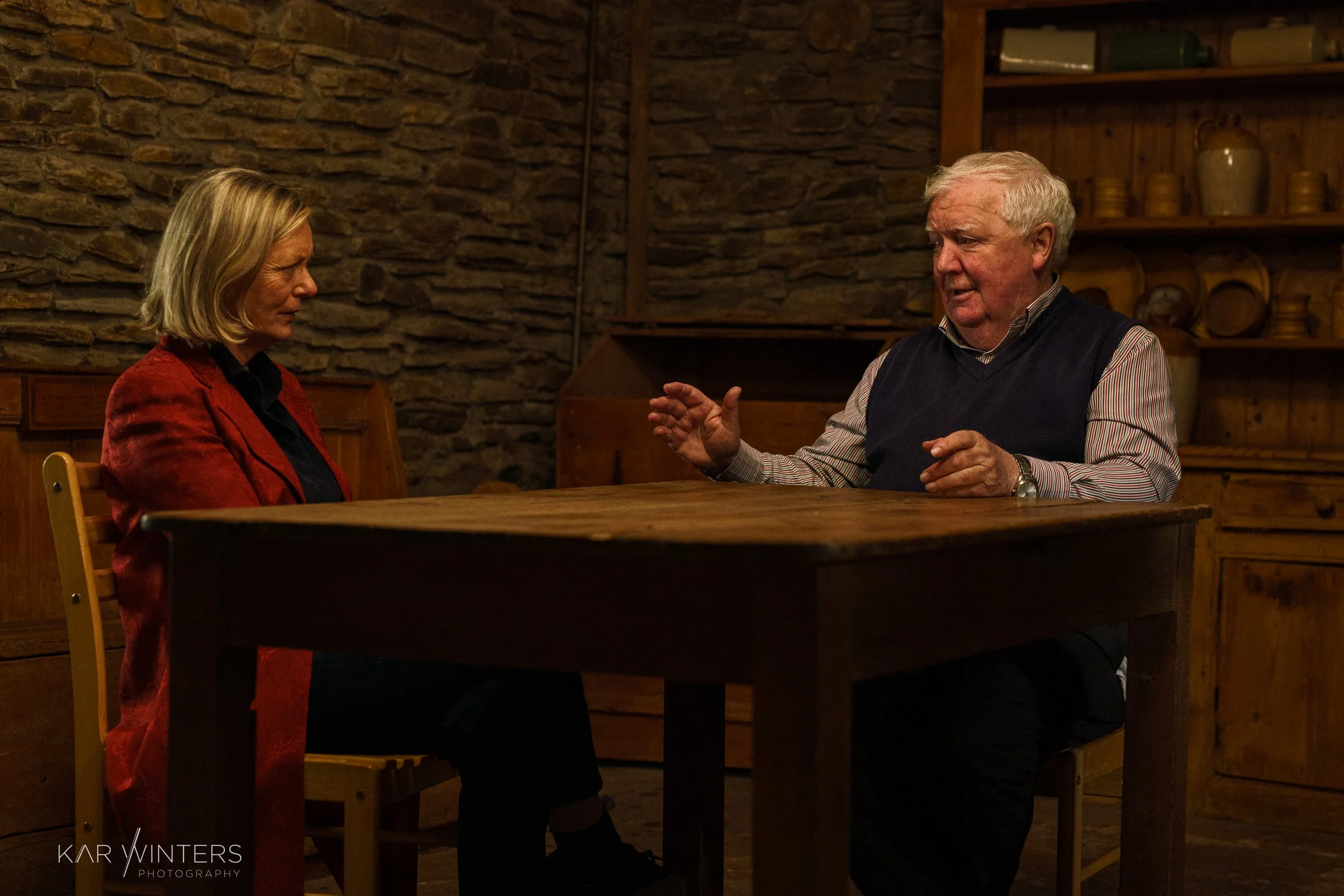 An older man and a woman are sitting across from each other at a wooden table, engaged in conversation inside a rustic room with stone walls and wooden shelves.