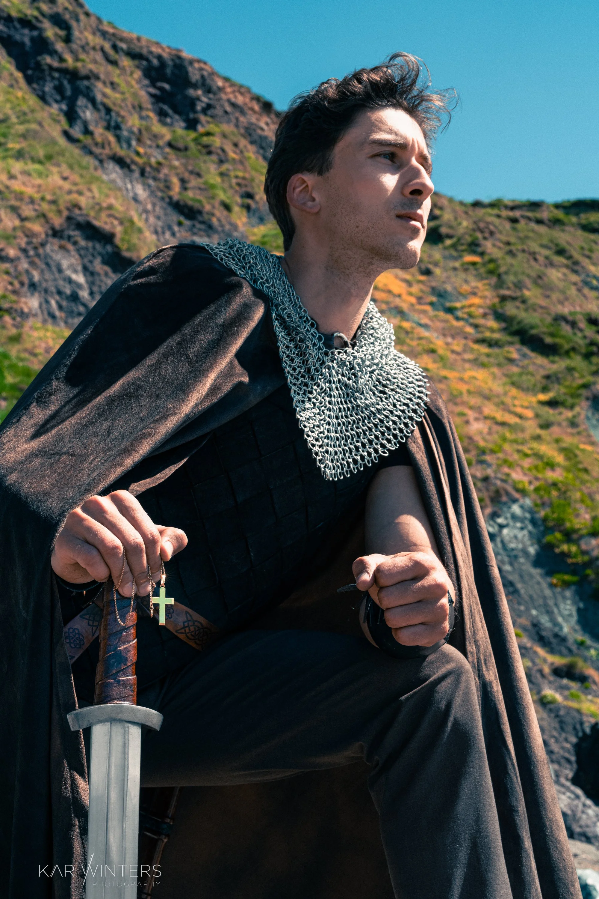 A man dressed as a knight, wearing chainmail and holding a sword, kneels on one knee outdoors with rocky hills and a blue sky in the background.