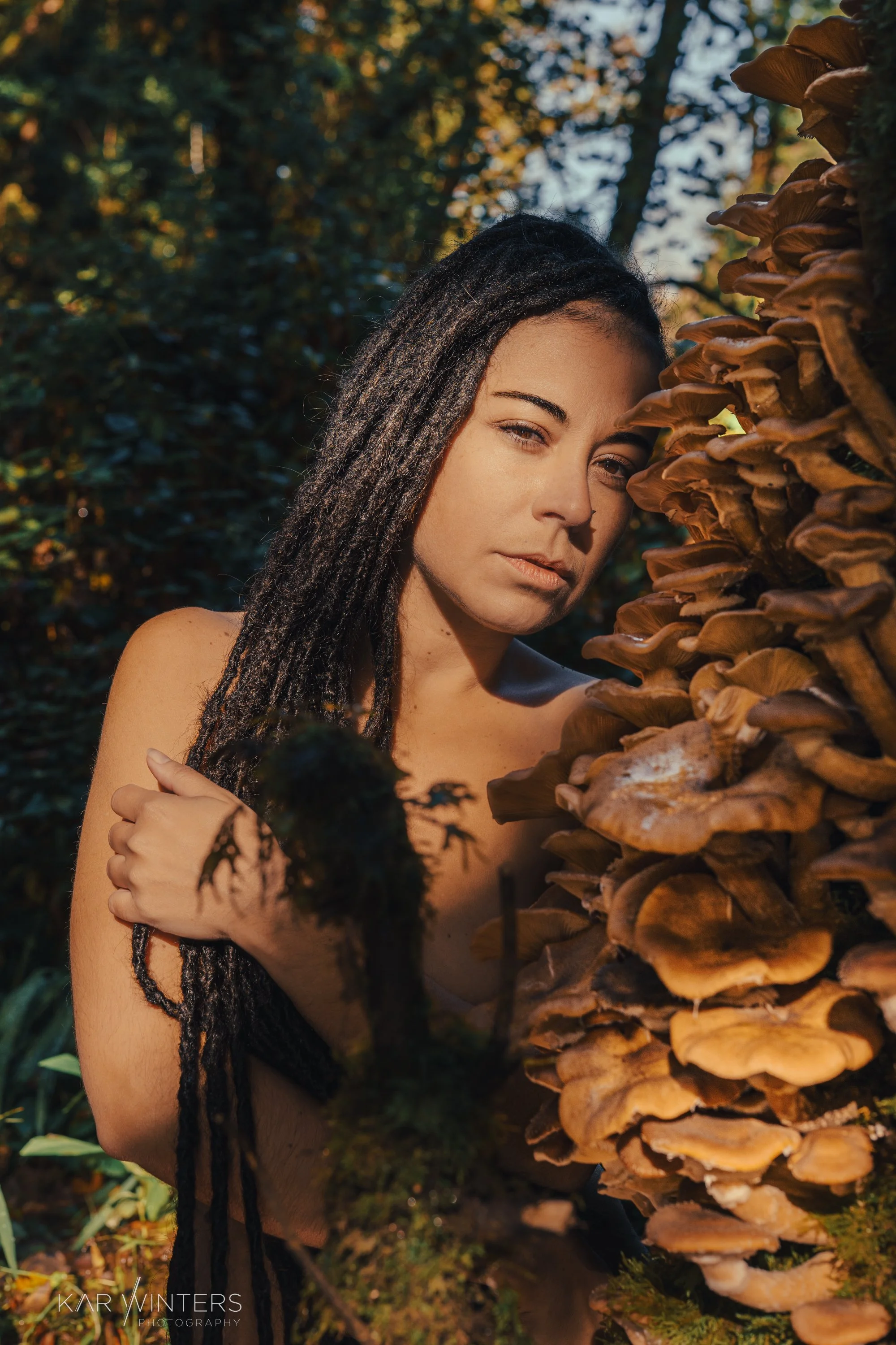 A woman with long dark dreadlocks posing outdoors next to a large cluster of brown mushrooms on a tree trunk, with a background of autumn foliage and trees.