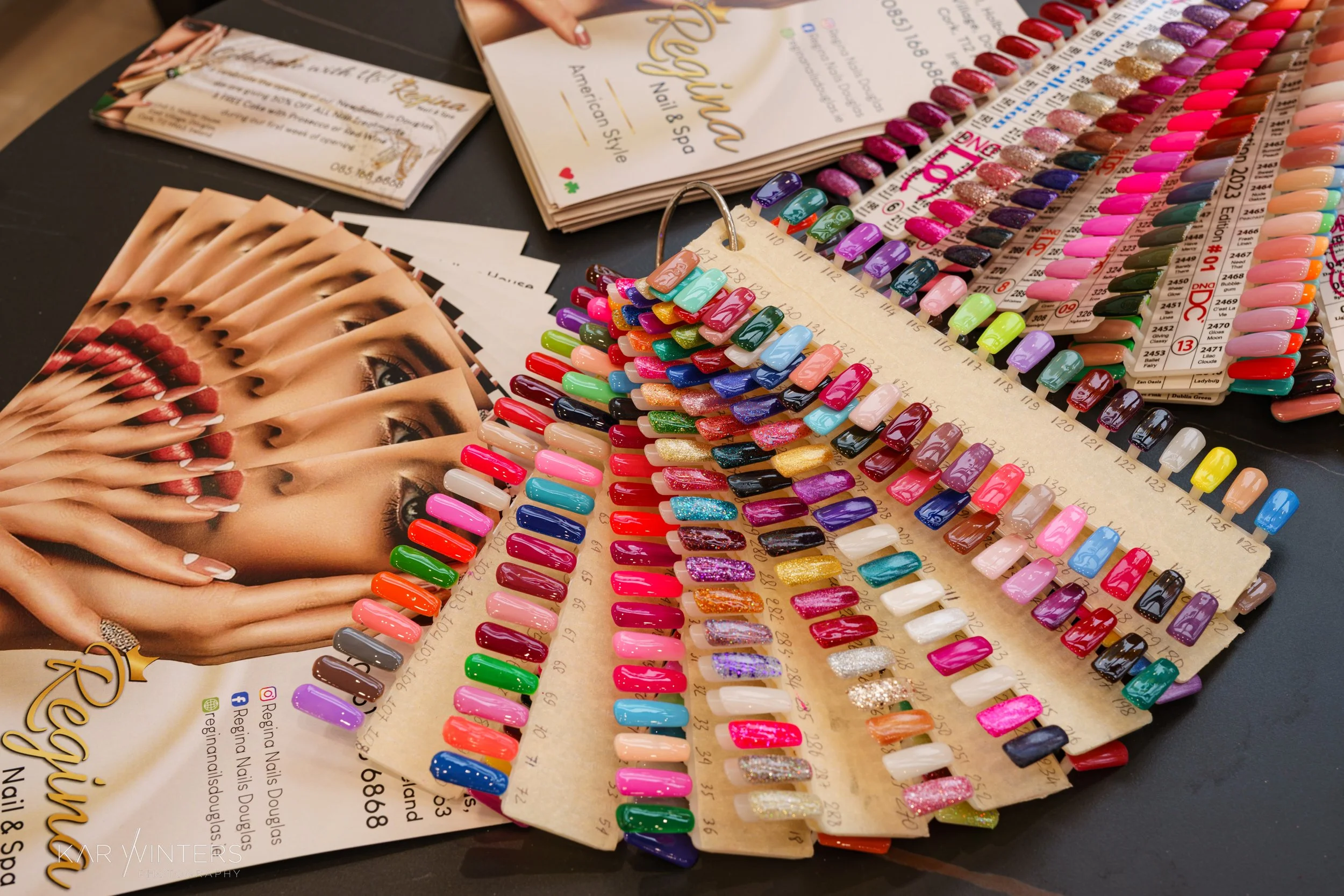 A display of various nail polish color samples arranged on color swatch boards, with a nail and spa brochure and a photo of a woman with red nails in the background.