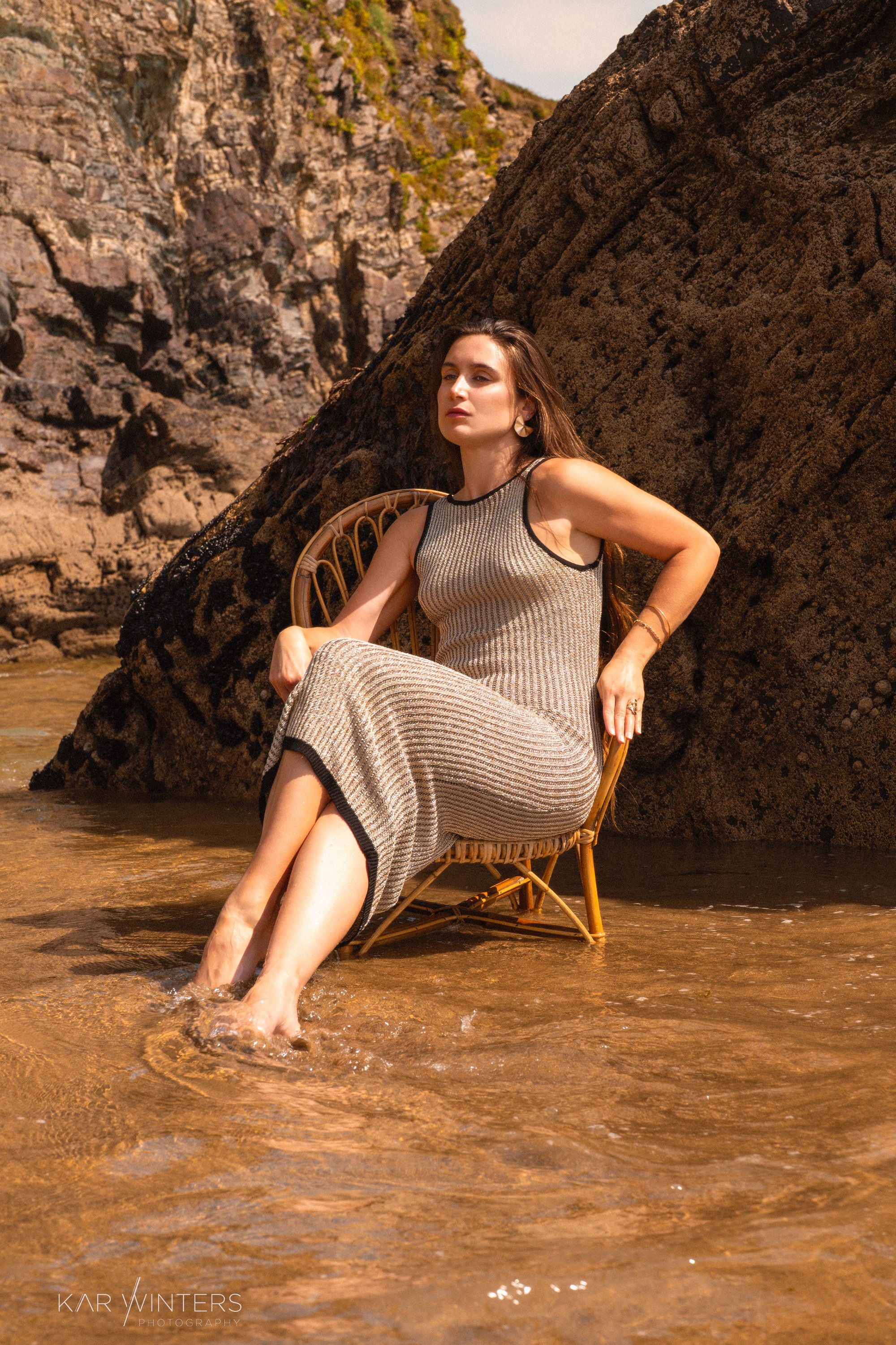 A woman in a sleeveless dress sitting on a wicker chair with her feet in shallow water, near large rocks and cliffs, outdoors.