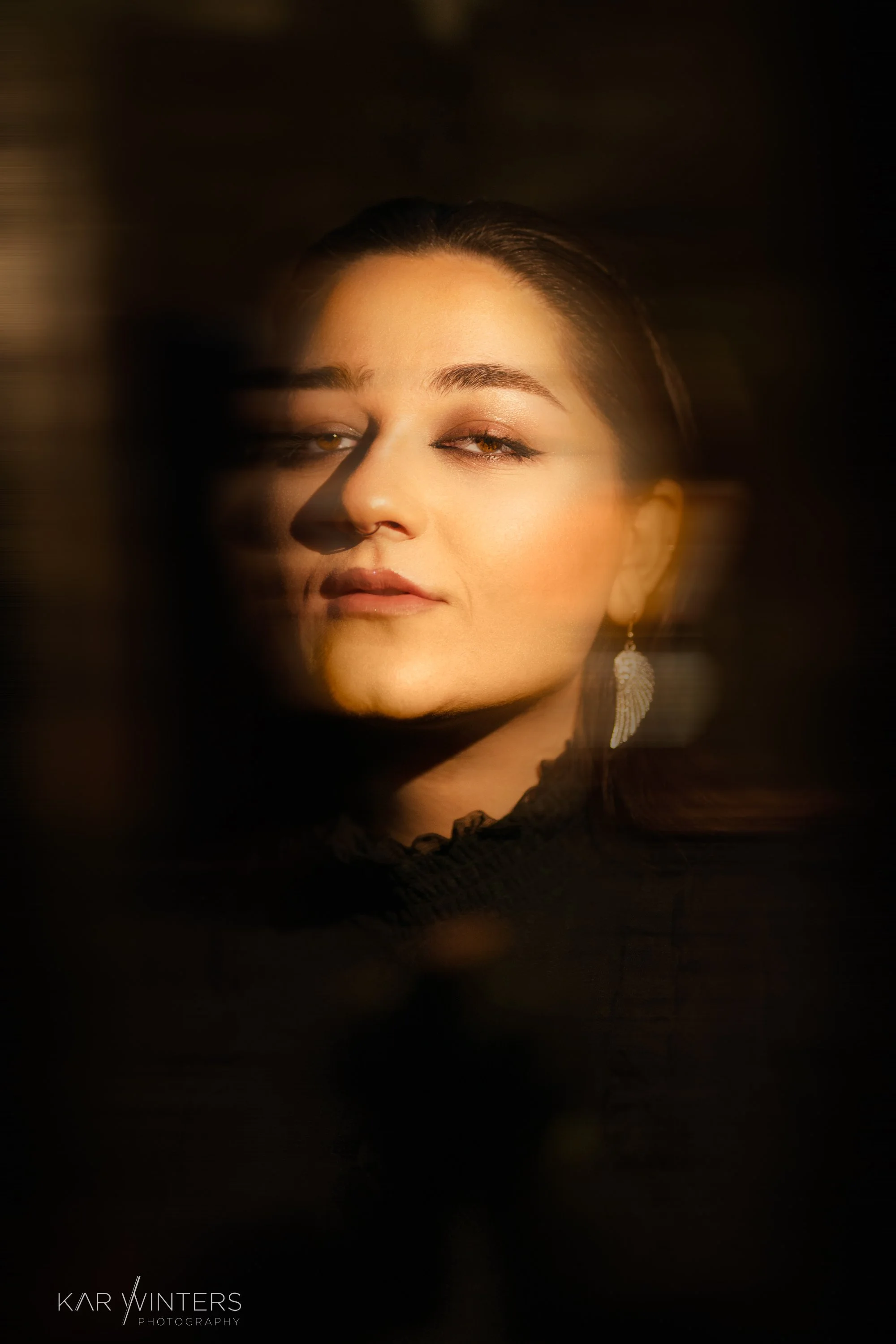 Close-up portrait of a woman with light skin, dark hair, one eye half-closed, wearing an earring, dark top, and looking at the camera with a confident expression. Photo by Kar Winters.