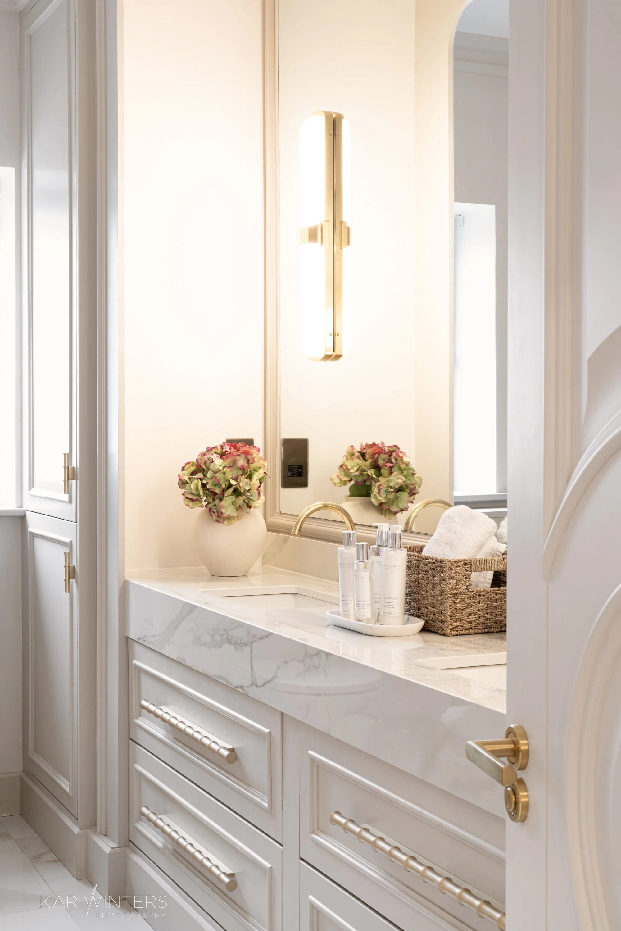 A bathroom vanity with a marble countertop, a mirror, and a wall-mounted light fixture. There are two vases with pink and green hydrangeas, a wicker basket with towels, and bottles of toiletries on a tray.