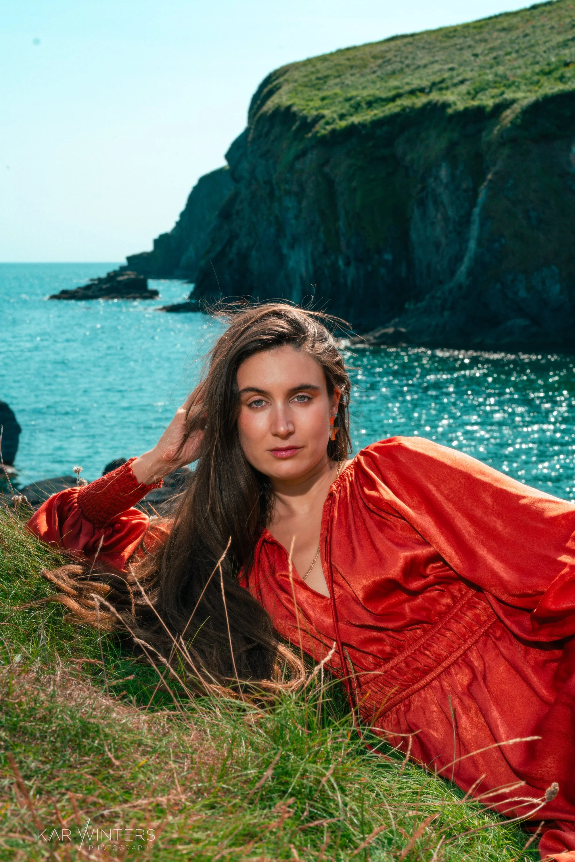 Woman lying on grass near a rocky shoreline with cliffs and ocean in the background, wearing a red satin dress.