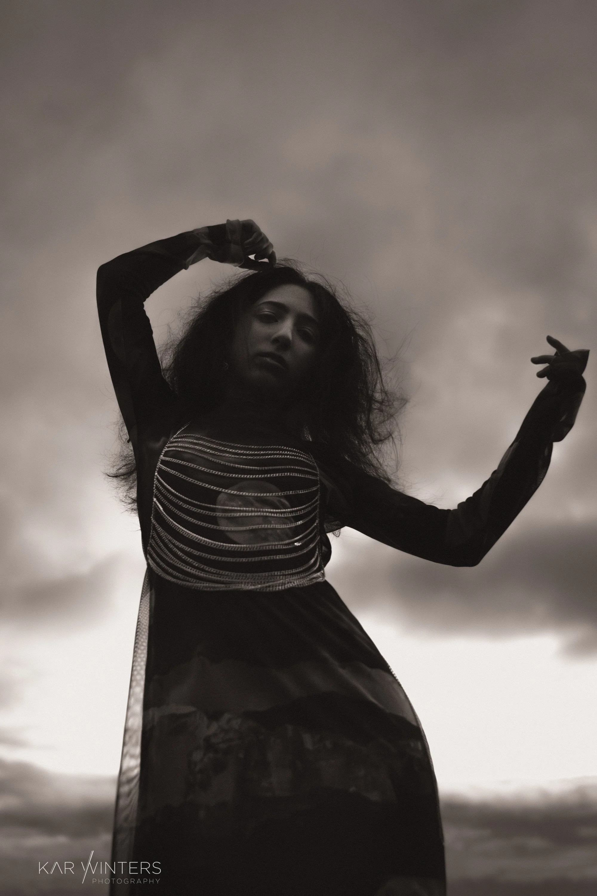 Black and white photo of a woman with curly hair, posing against a cloudy sky. She is wearing a long, dark dress with sheer sleeves and a striped bodice. Her arms are raised, with one hand touching her head and the other extended outward.