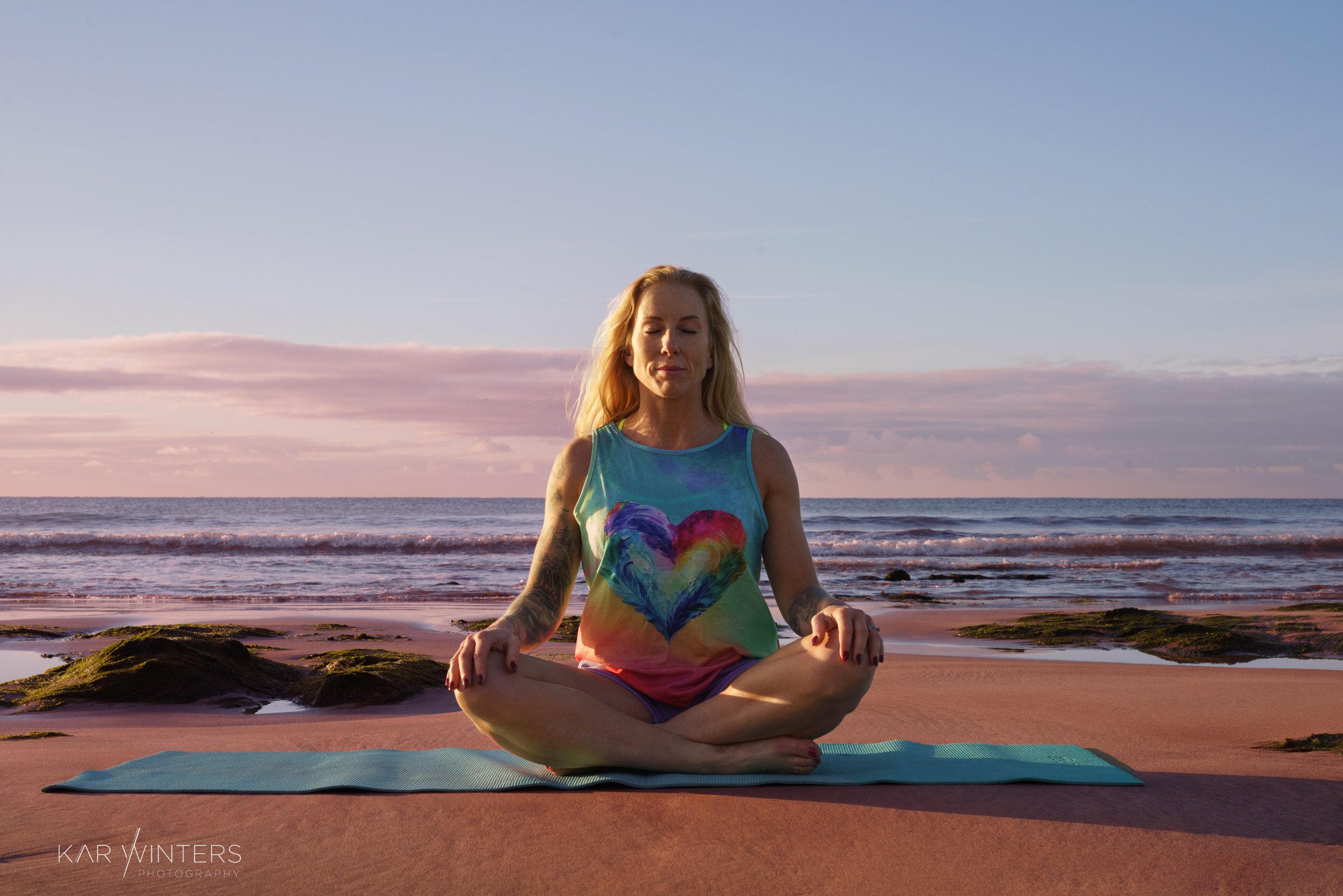 A woman practicing yoga on a surfboard on a beach during sunrise or sunset, with rocks and the ocean in the background.
