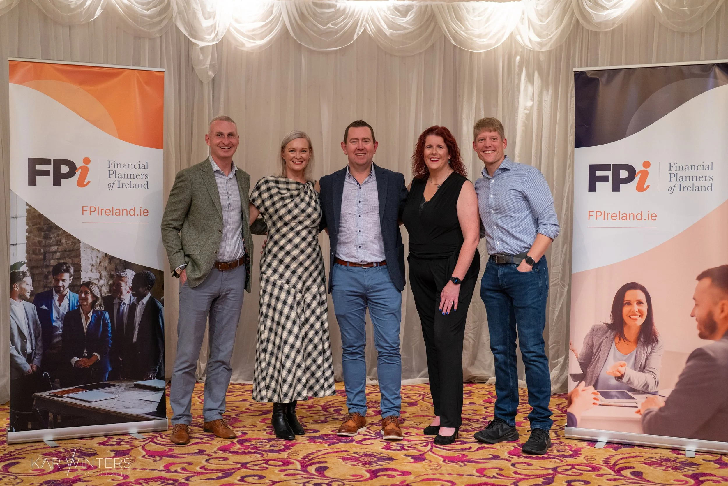 Group of five people, three men and two women, standing together and smiling in front of two banners for Financial Planners of Ireland, inside a decorated room with curtains and patterned carpet.