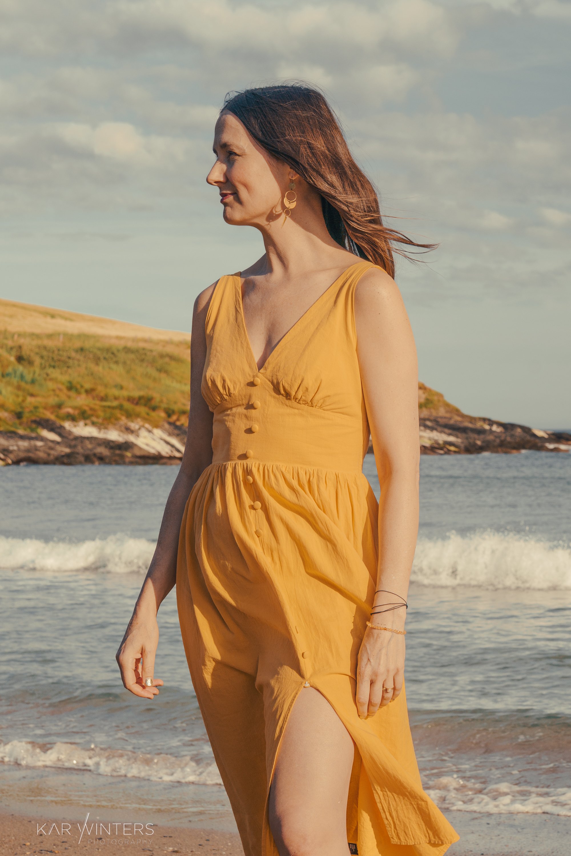 Woman in a yellow dress standing by the shoreline, looking to the side, with waves and a hilly landscape in the background.
