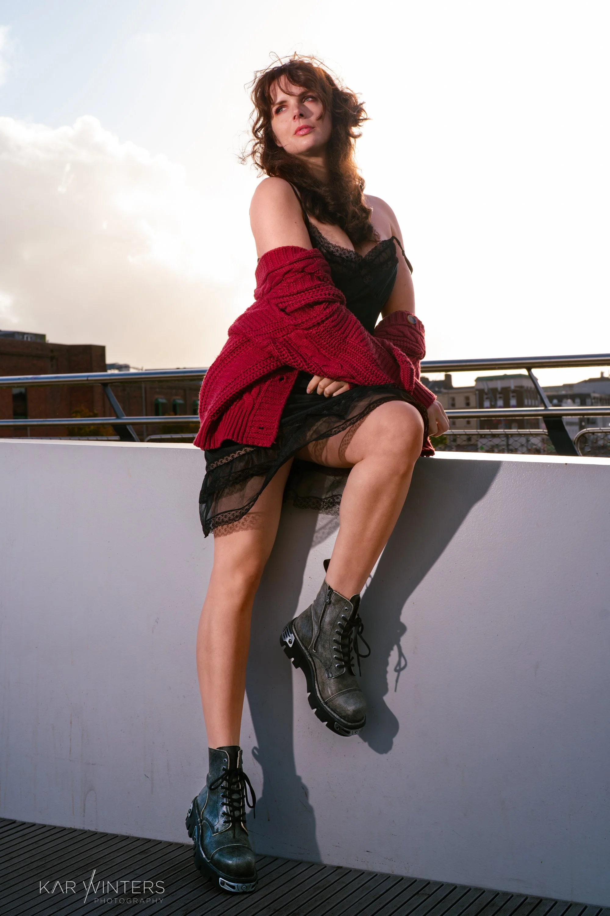 A woman sitting on a rooftop ledge, wearing black lace clothing, a red knitted cardigan, and black combat boots, with city buildings in the background at sunset.