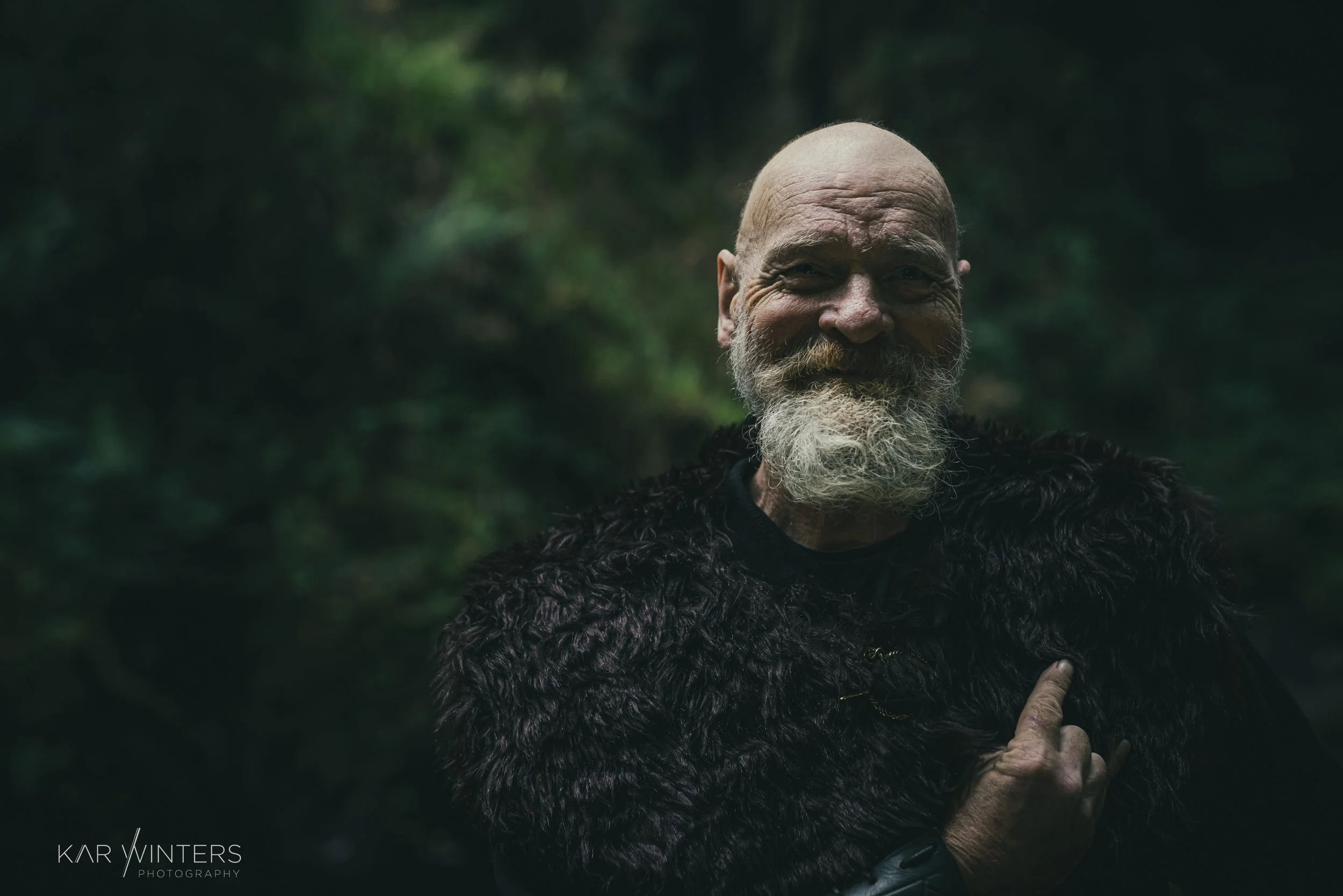 An elderly man with a white beard and mustache wearing a dark, textured fur-like coat, standing outdoors in a forest with blurred green foliage in the background.