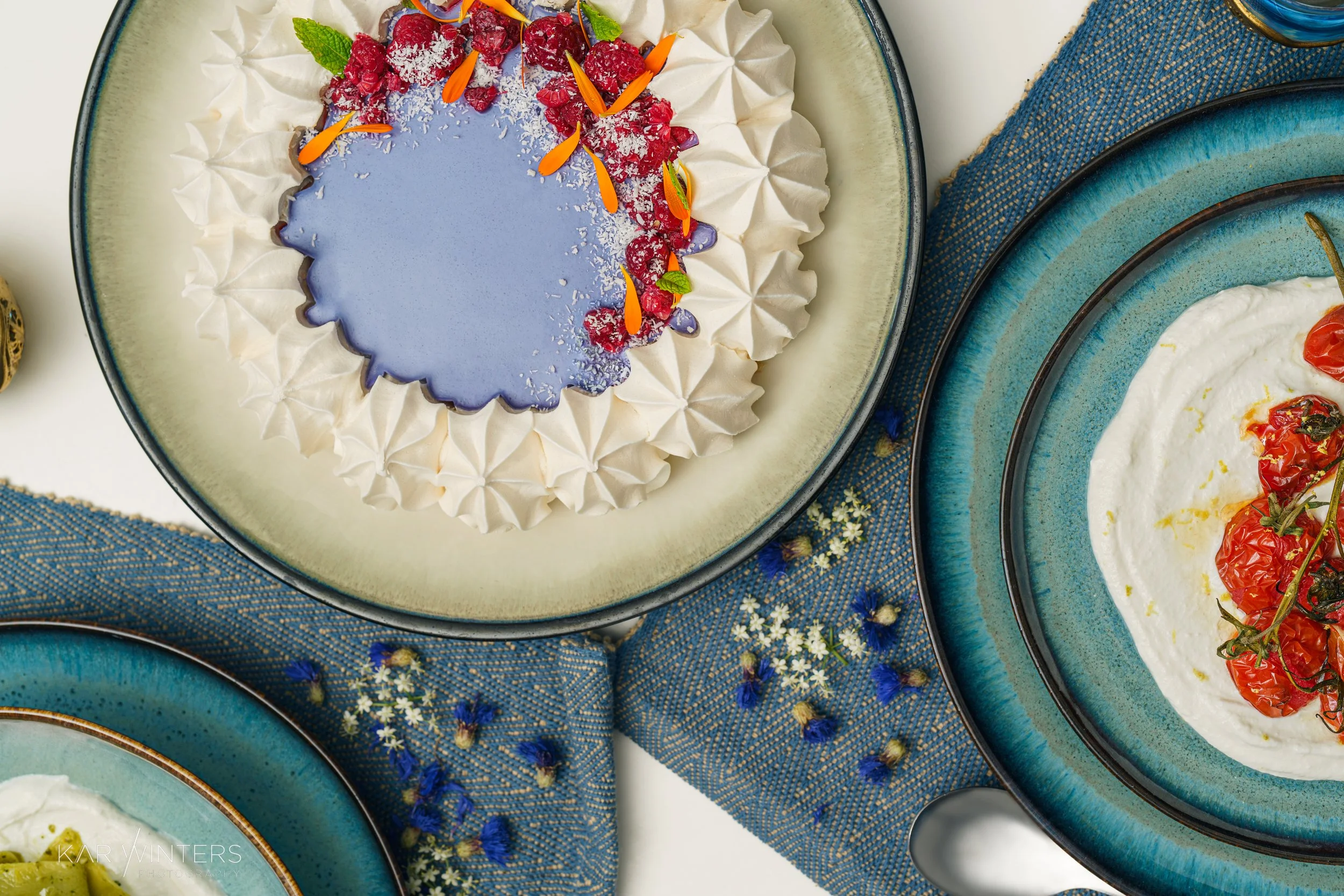 Assorted plates of gourmet food, including a decorated cake with purple icing, raspberries, and edible flowers; a plate with roasted tomatoes in a creamy sauce; and another with sliced green vegetables, on blue-patterned napkins.