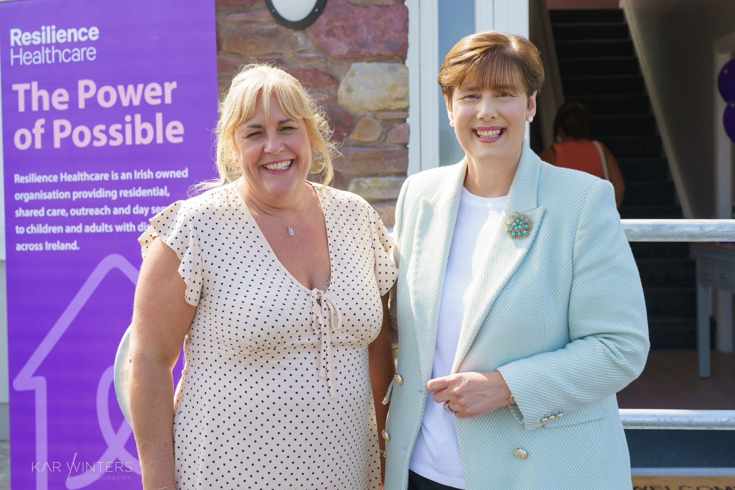Two women smiling standing outdoors in front of a purple sign that reads 'Resilience Healthcare. The Power of Possible,' with a brief description of the organization.