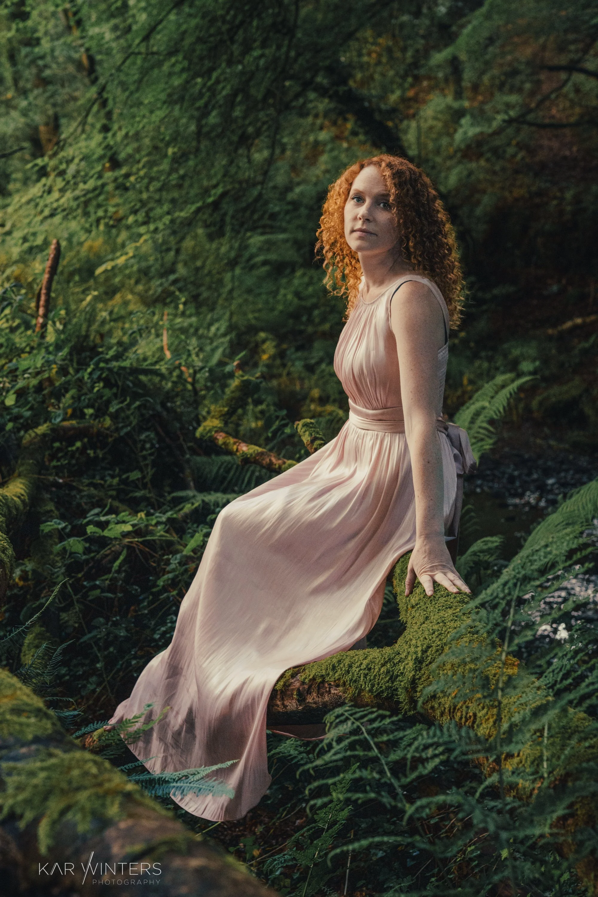 A woman with curly red hair in a flowing beige dress sitting on a moss-covered log in a dense forest.