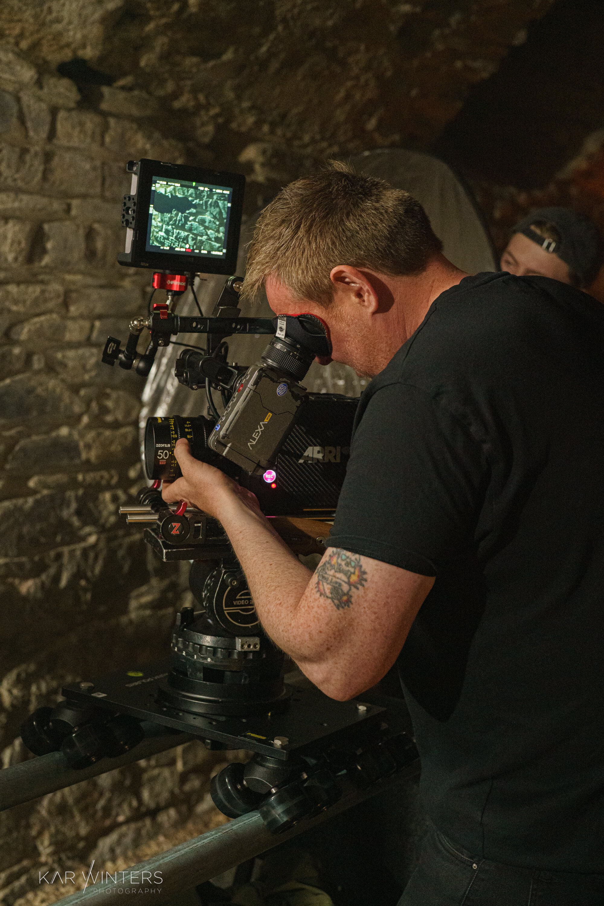 A person with a tattoo on their arm operating a professional camera on a rig, filming in a dimly lit environment with stone walls.