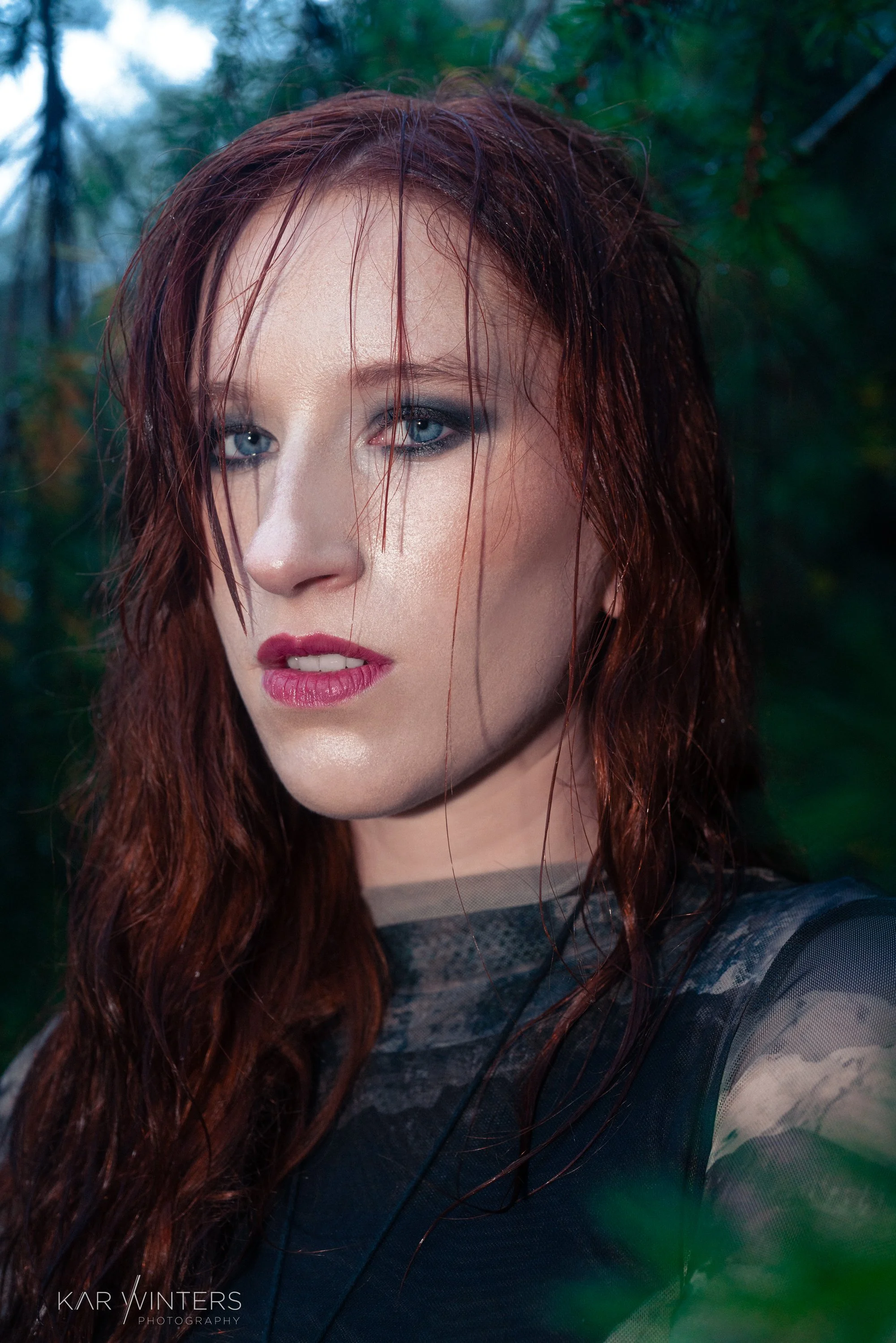 Close-up portrait of a woman with wet red hair and blue eyes, standing outdoors with green foliage in the background.