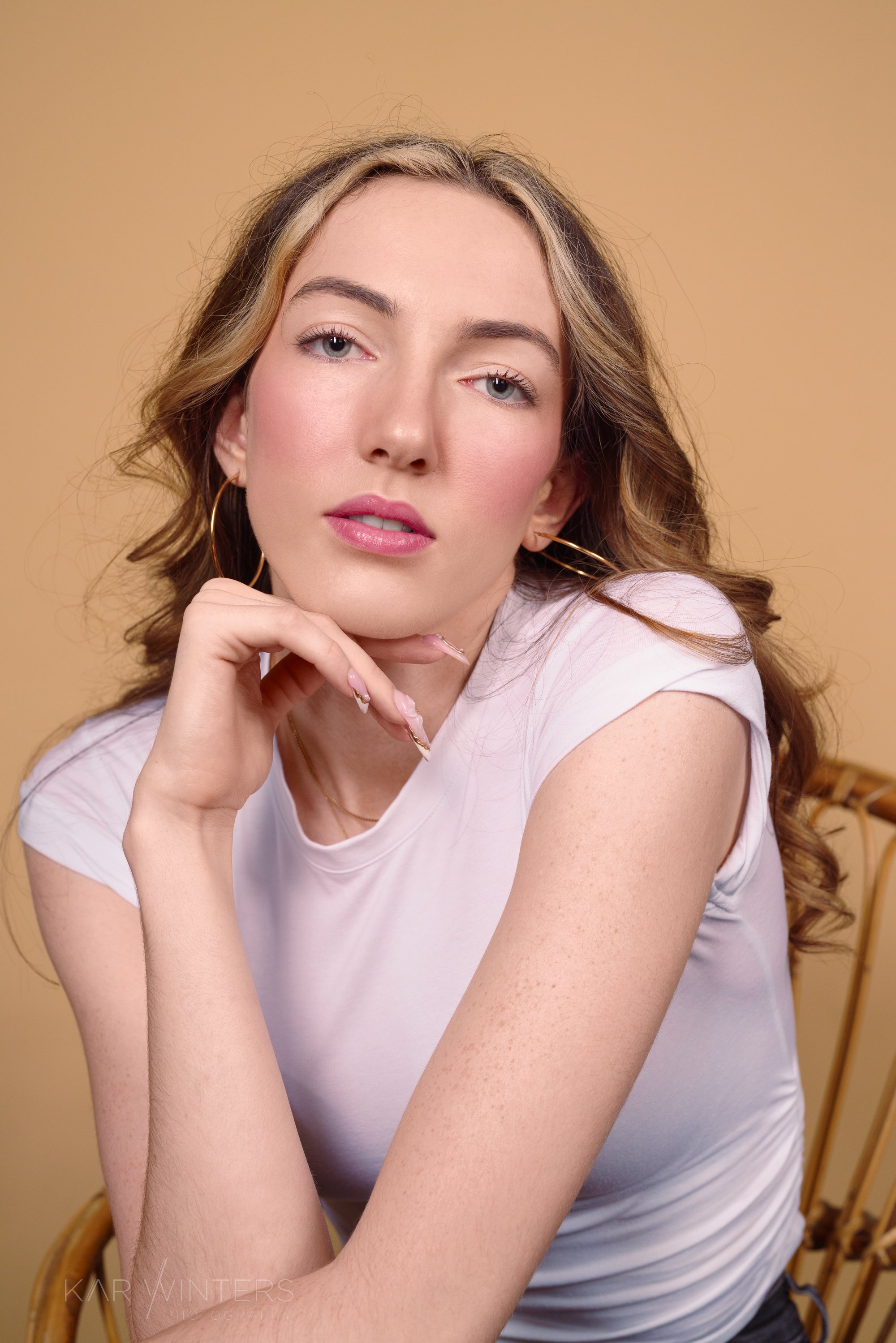 Close-up portrait of a young woman with blue eyes, wavy light brown hair with blonde highlights, wearing gold hoop earrings, a white t-shirt, resting her chin on her hand, looking slightly to the camera, with a neutral beige background.