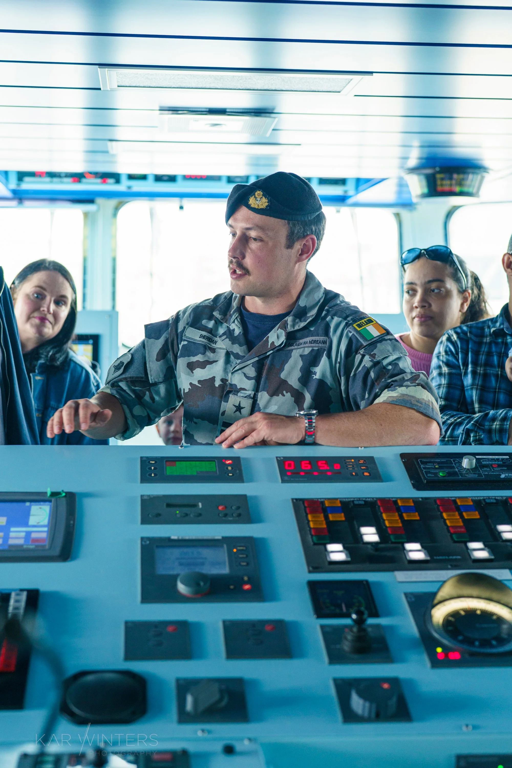 A group of people, including a man in a military uniform, are gathered on a ship's bridge, operating navigation and control equipment.