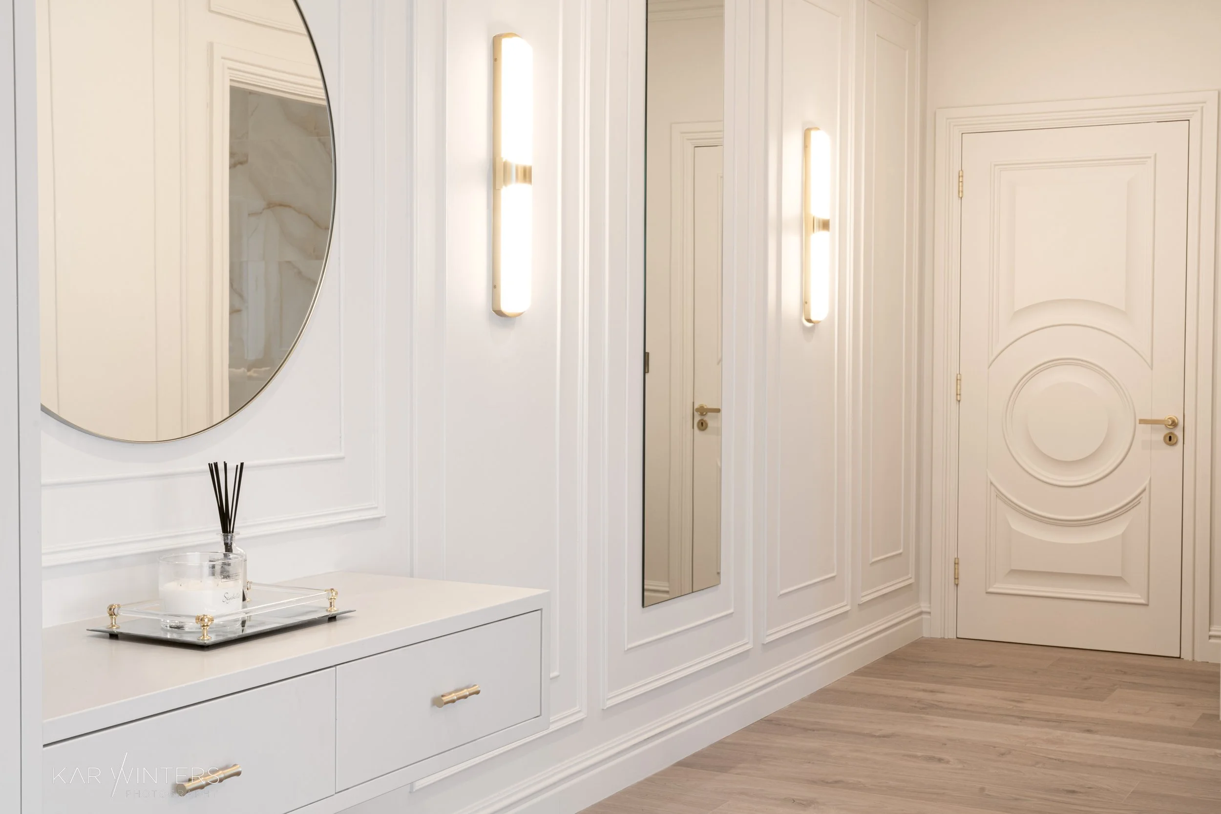 Stylish white entryway with dual vertical wall-mounted lights, a large oval mirror, a small white console table with a tray holding candles and reed diffuser, decorative molding, a mirror, and a door with circular panel detail.