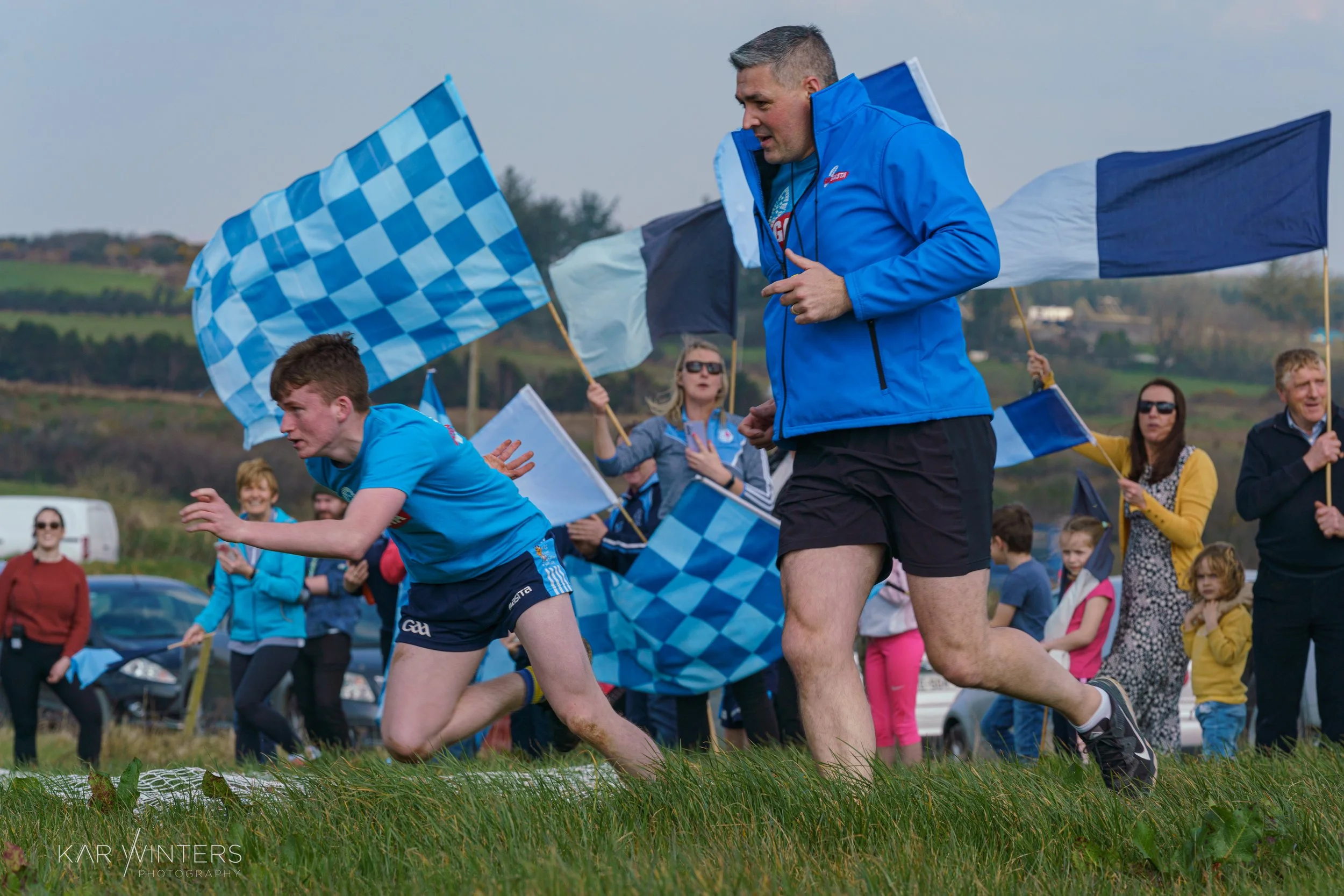 A young athlete in blue shorts and a blue jersey sprints across a grassy field while an older man, also in blue, runs alongside him. A crowd of spectators, some holding blue and white checkered flags, watch in the background during a sports event out