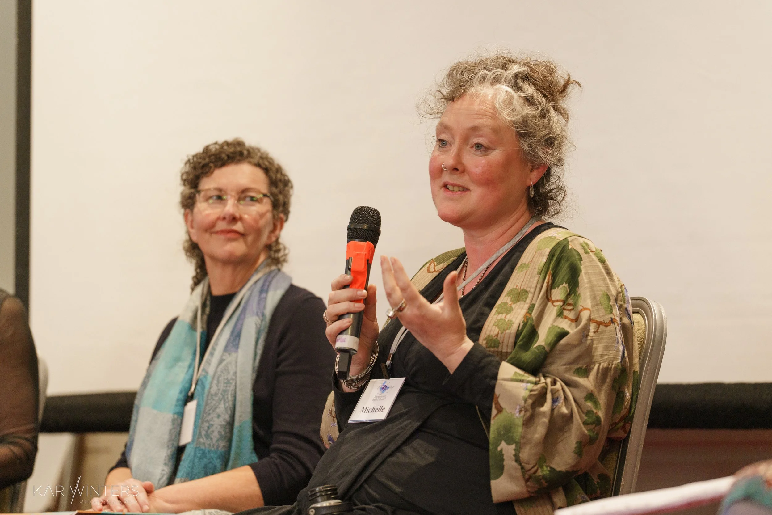 Two women sitting on a panel at a conference. The woman on the right, with curly gray hair, is speaking into a microphone and wearing a patterned jacket. The woman on the left, with short curly hair and glasses, is attentively listening.