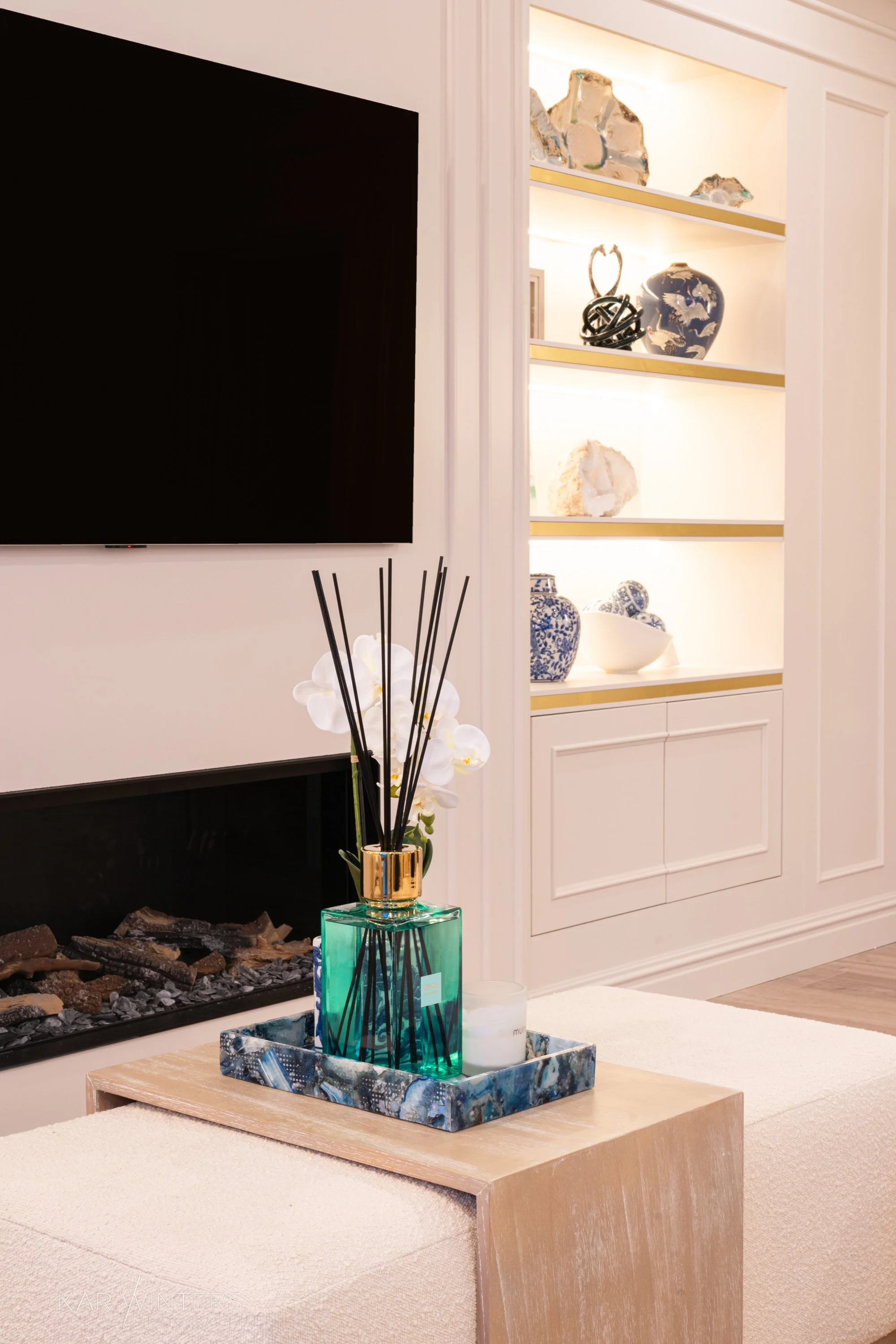 Living room with a wall-mounted television, a decorated fireplace, a wooden coffee table with a diffuser and a candle, and built-in white shelves with decorative vases and objects.