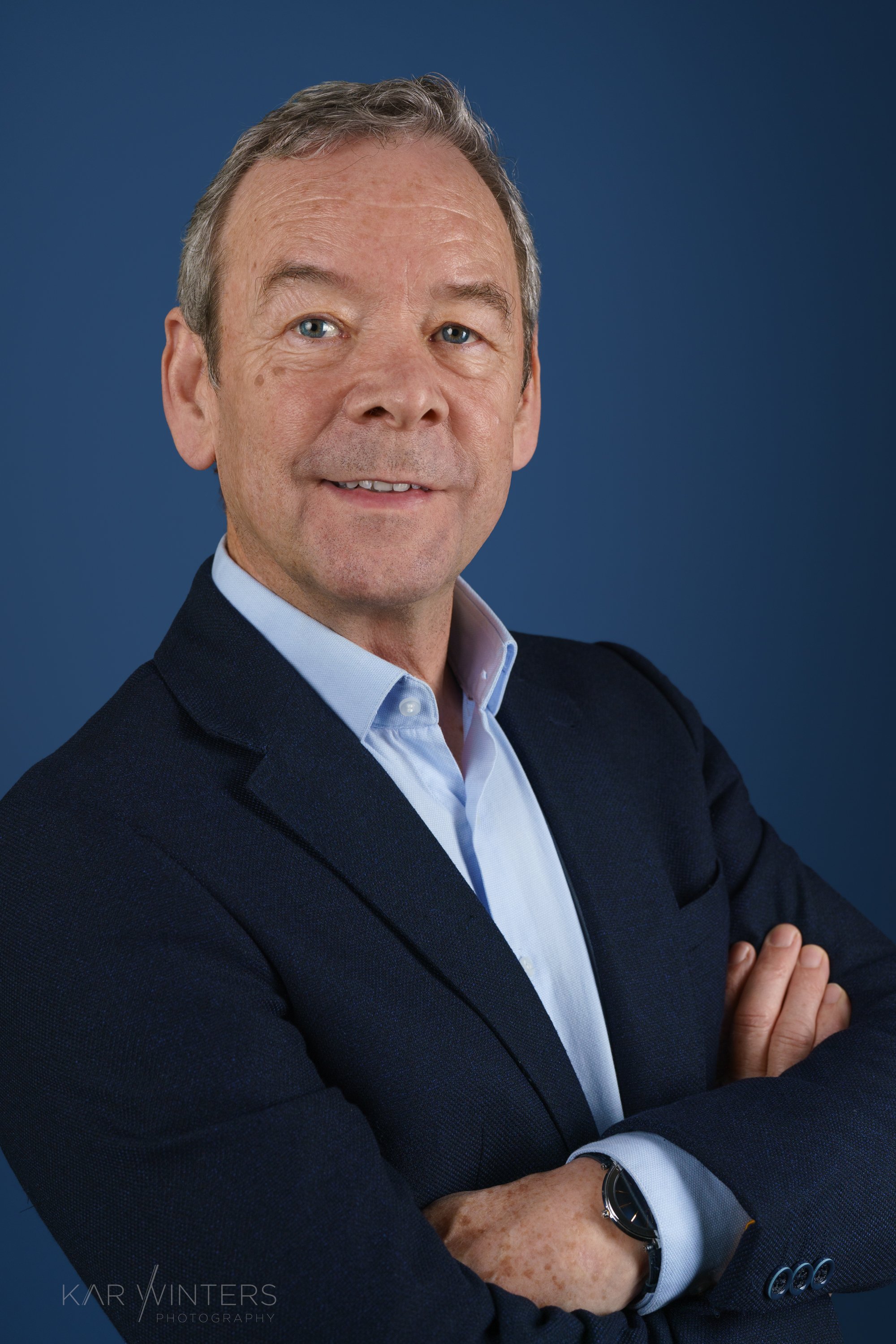 A professional middle-aged man in a dark blue blazer and light blue shirt with crossed arms posing against a solid dark blue background.