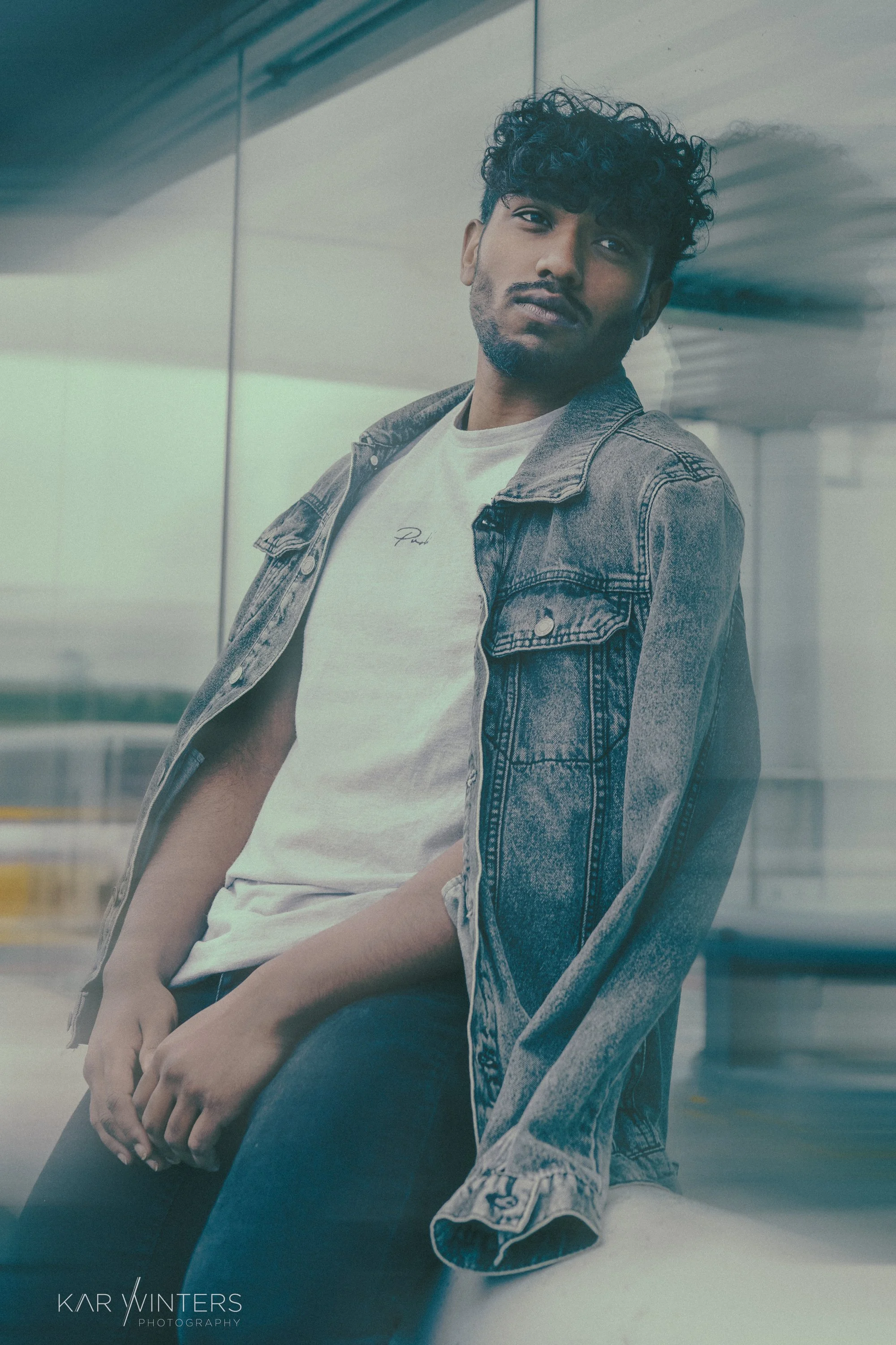 A young man with curly hair and a beard wearing a white t-shirt and denim jacket, leaning against a reflective wall at an airport or transportation terminal.