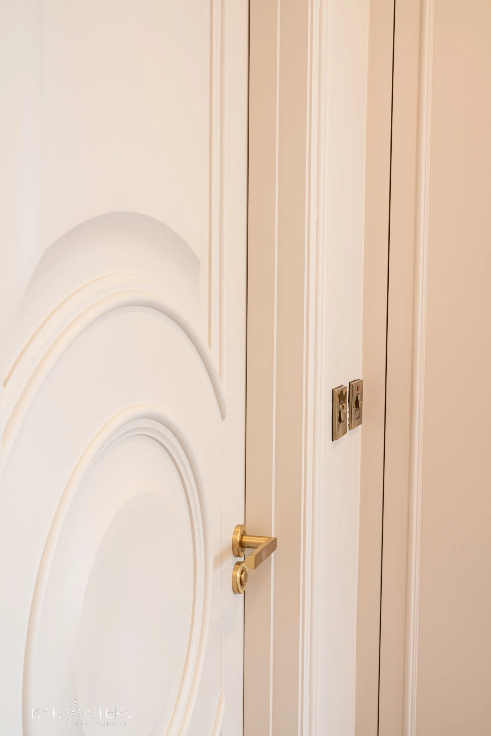 Close-up view of a white interior door with decorative moldings, gold door handle, and a door lock. Part of a white wall and a closet door with two metallic handle plates are visible.