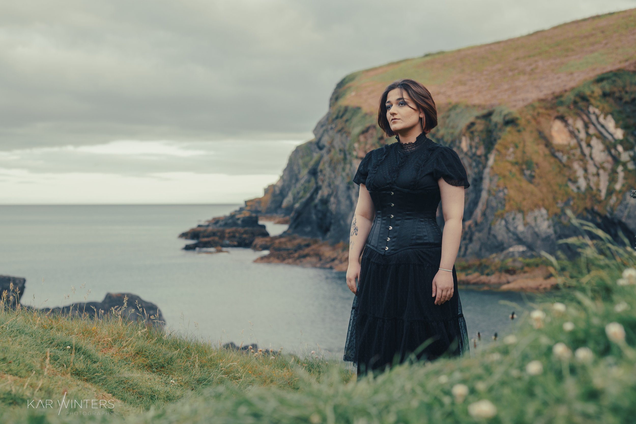 A woman with short brown hair in a black dress standing on a grassy hill near a coastal cliff with the ocean in the background.