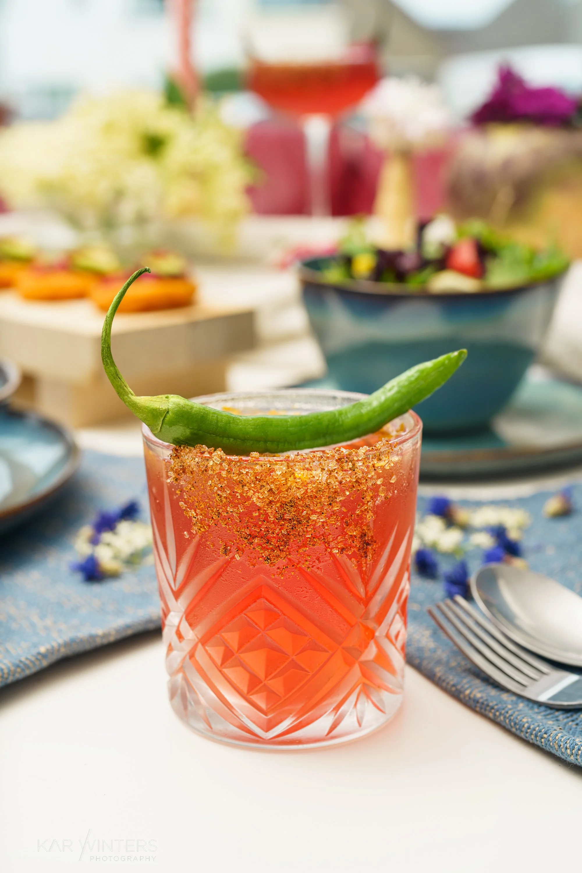 A pink cocktail garnished with a green chili on a white table, with a blue bowl of salad and other food and drinks in the background.