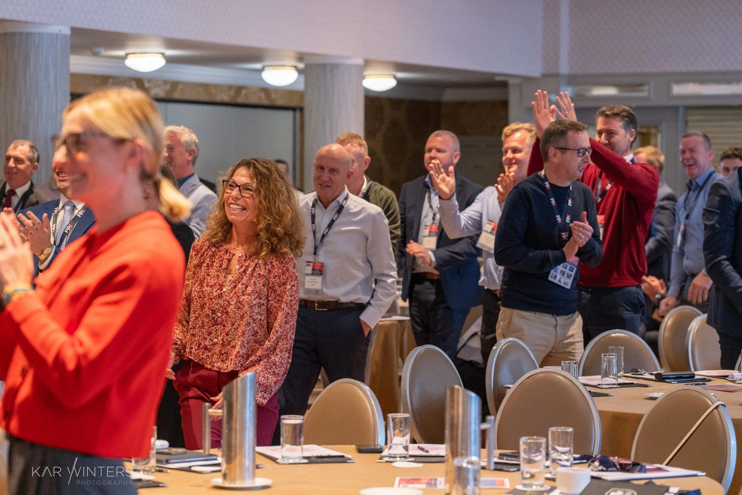 Group of businesspeople standing and clapping in a conference room, some smiling and engaging with each other.