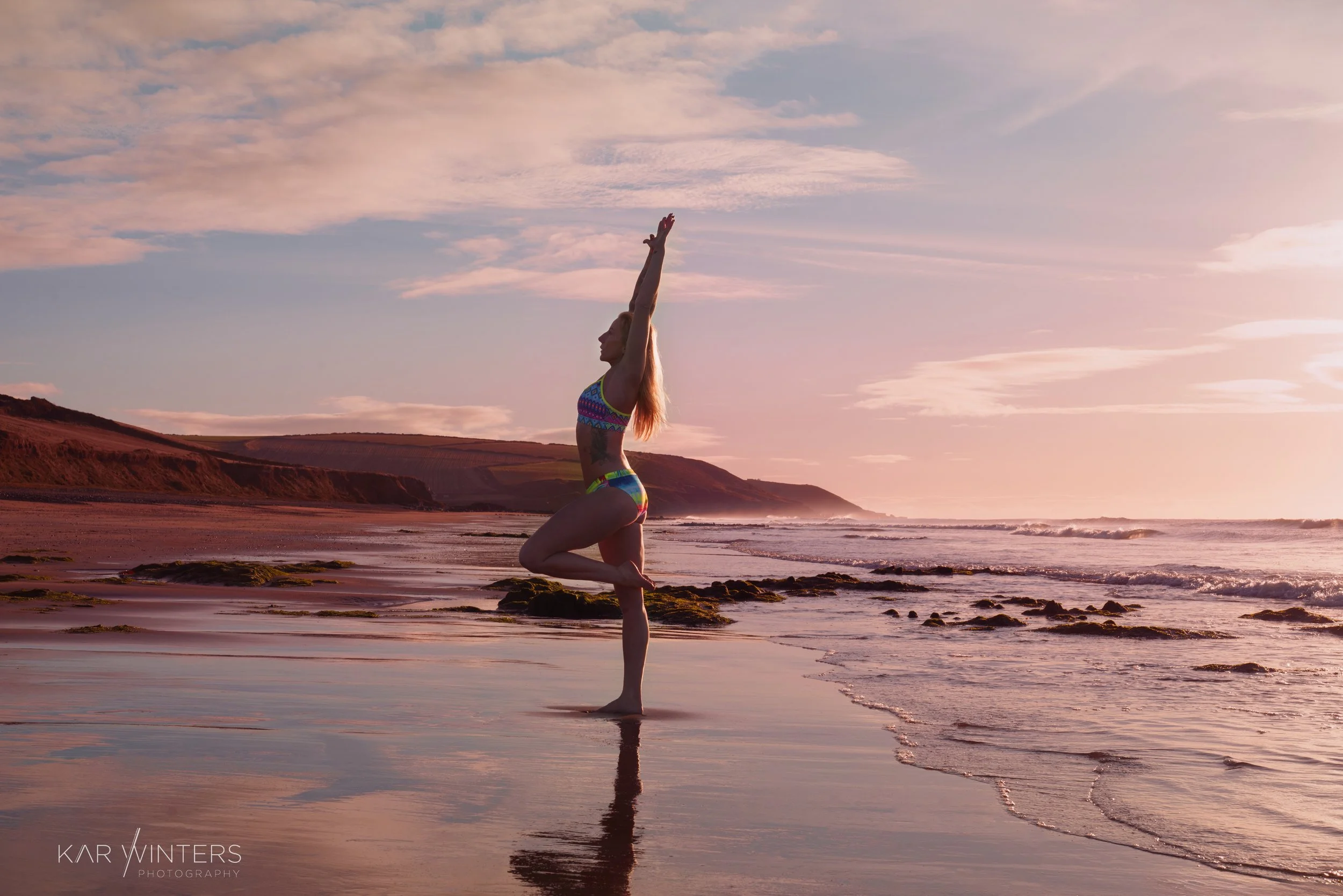 A woman practicing yoga on a beach at sunset, standing in a tree pose with arms raised overhead, overlooking the ocean and distant cliffs.