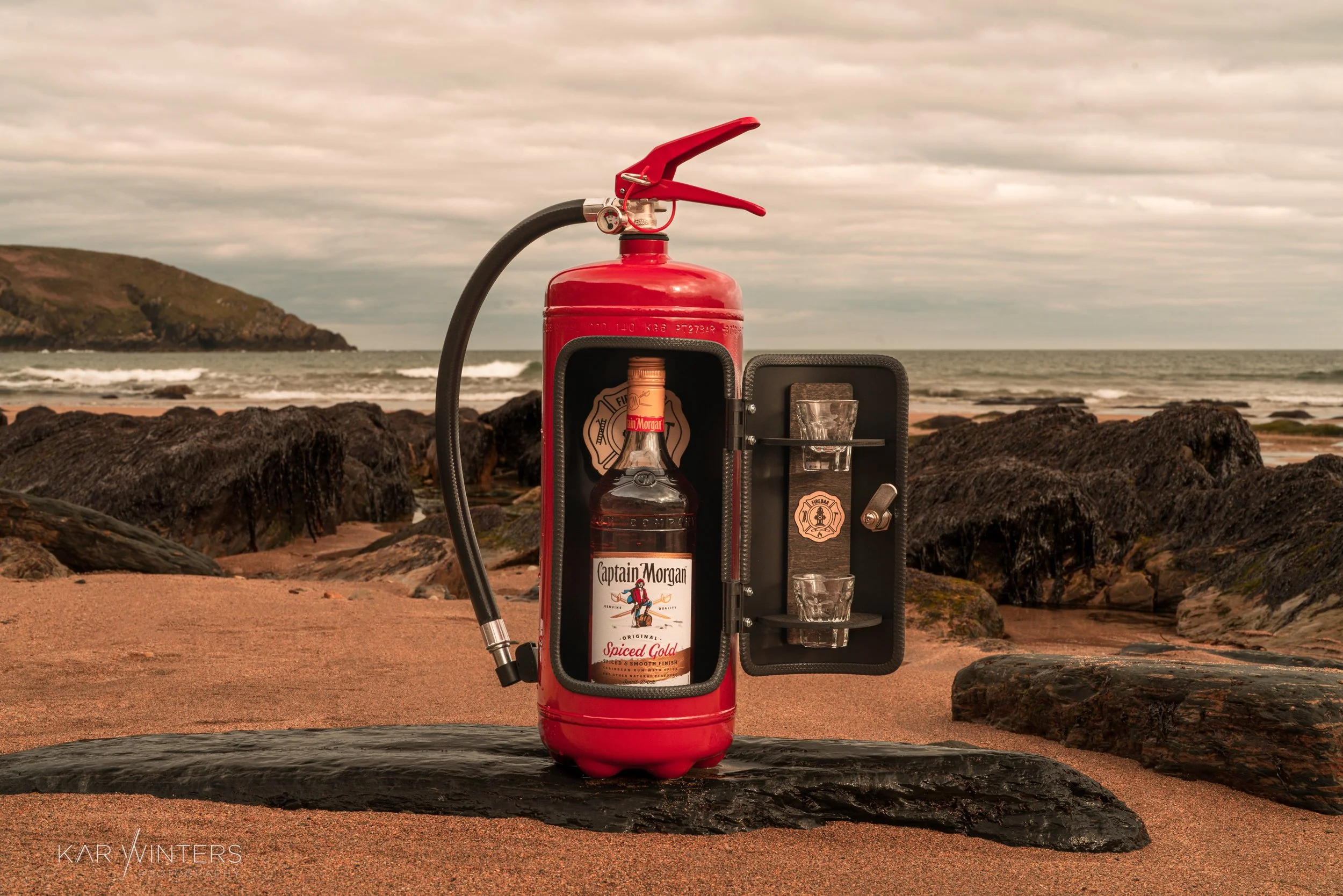 A fire extinguisher repurposed as a bar with a bottle of Captain Morgan Spiced Gold and shot glasses inside, placed on a rock on a beach with rocks and ocean in the background.