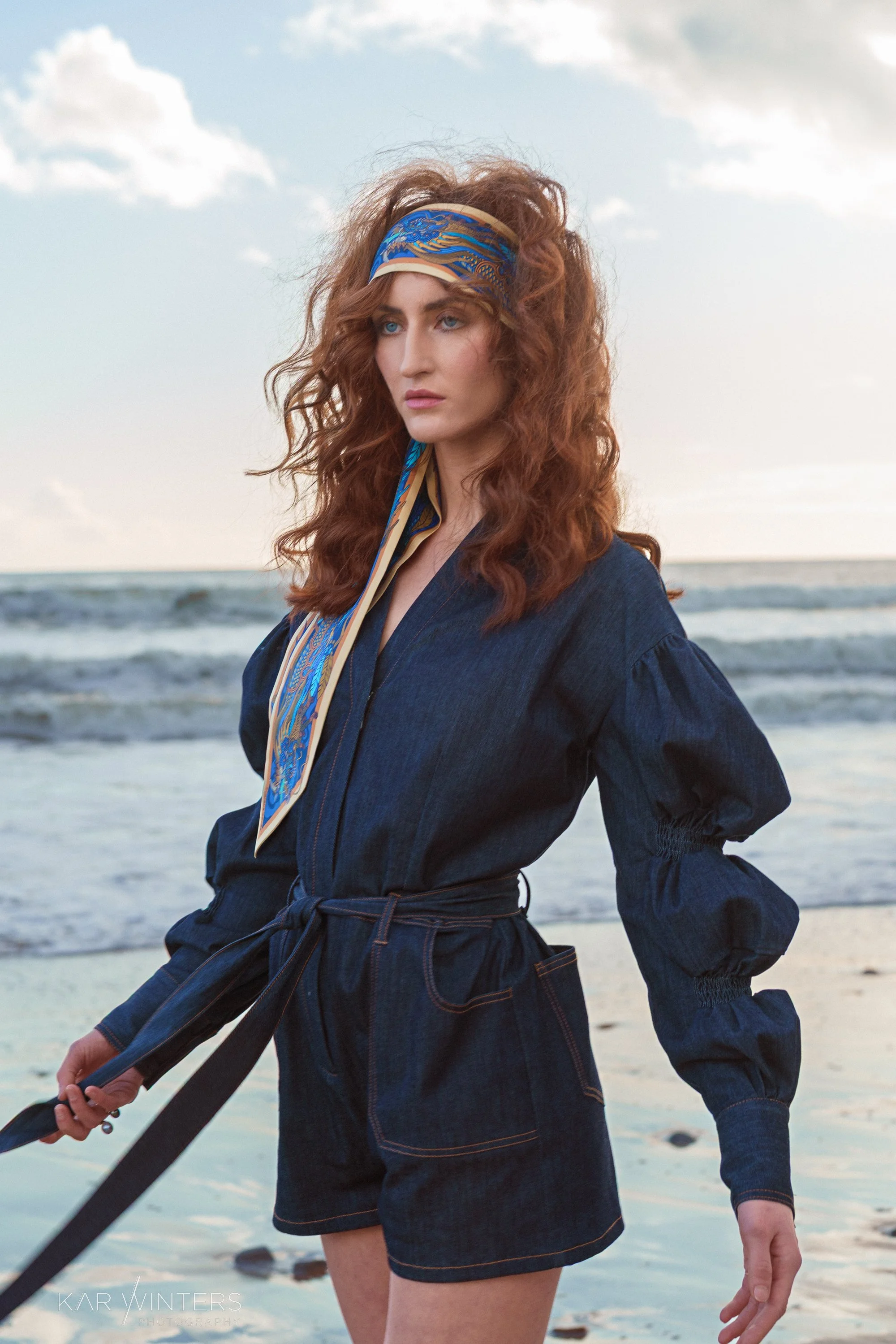 A woman with curly red hair and a blue patterned headscarf standing on a beach, wearing a denim jumpsuit with rolled-up shorts and long sleeves.