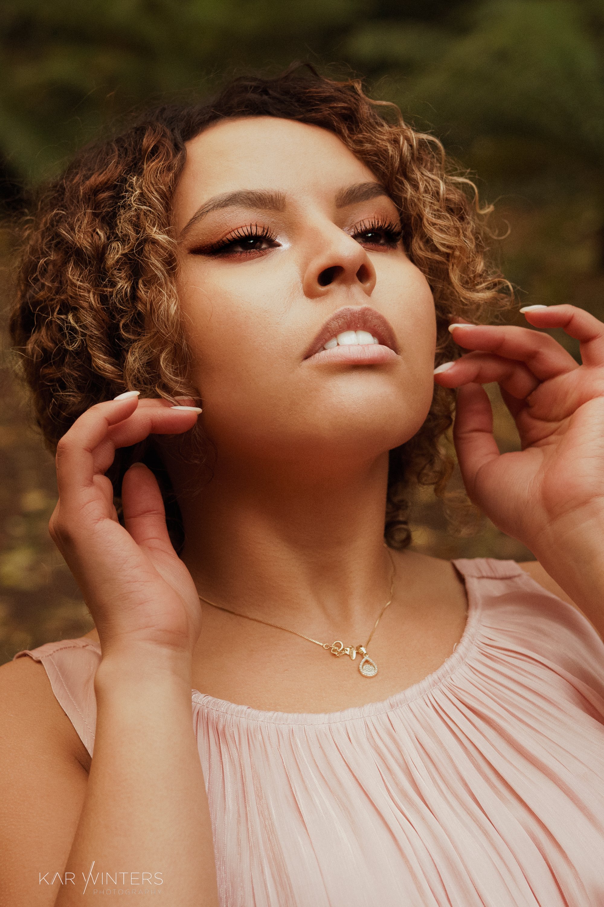 A woman with curly hair and light makeup posing outdoors, wearing a pink pleated top and a gold necklace.