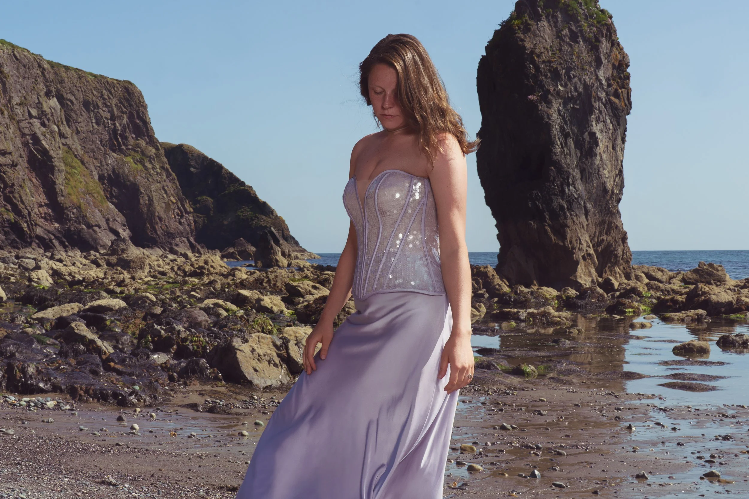 A woman in a strapless, silver dress standing on a rocky beach with large rock formations and the ocean in the background.