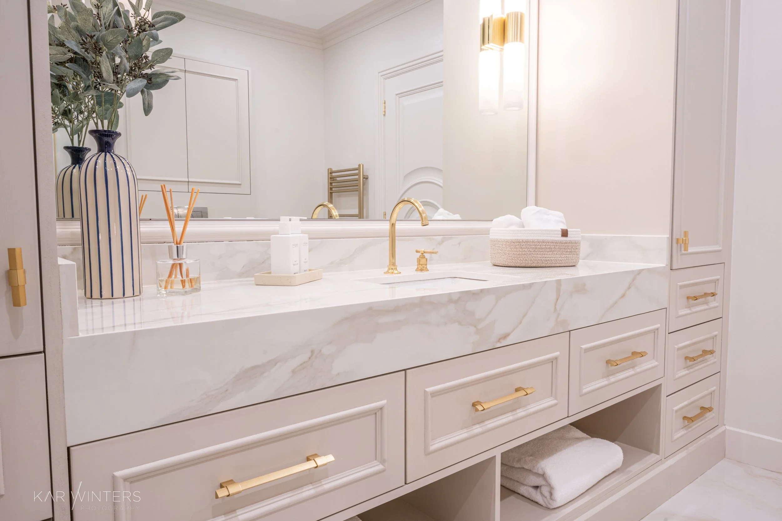 A luxurious bathroom vanity with a marble countertop, gold fixtures, decorative vases, and neatly rolled towels, with a large mirror reflecting a white wall and a lamp.