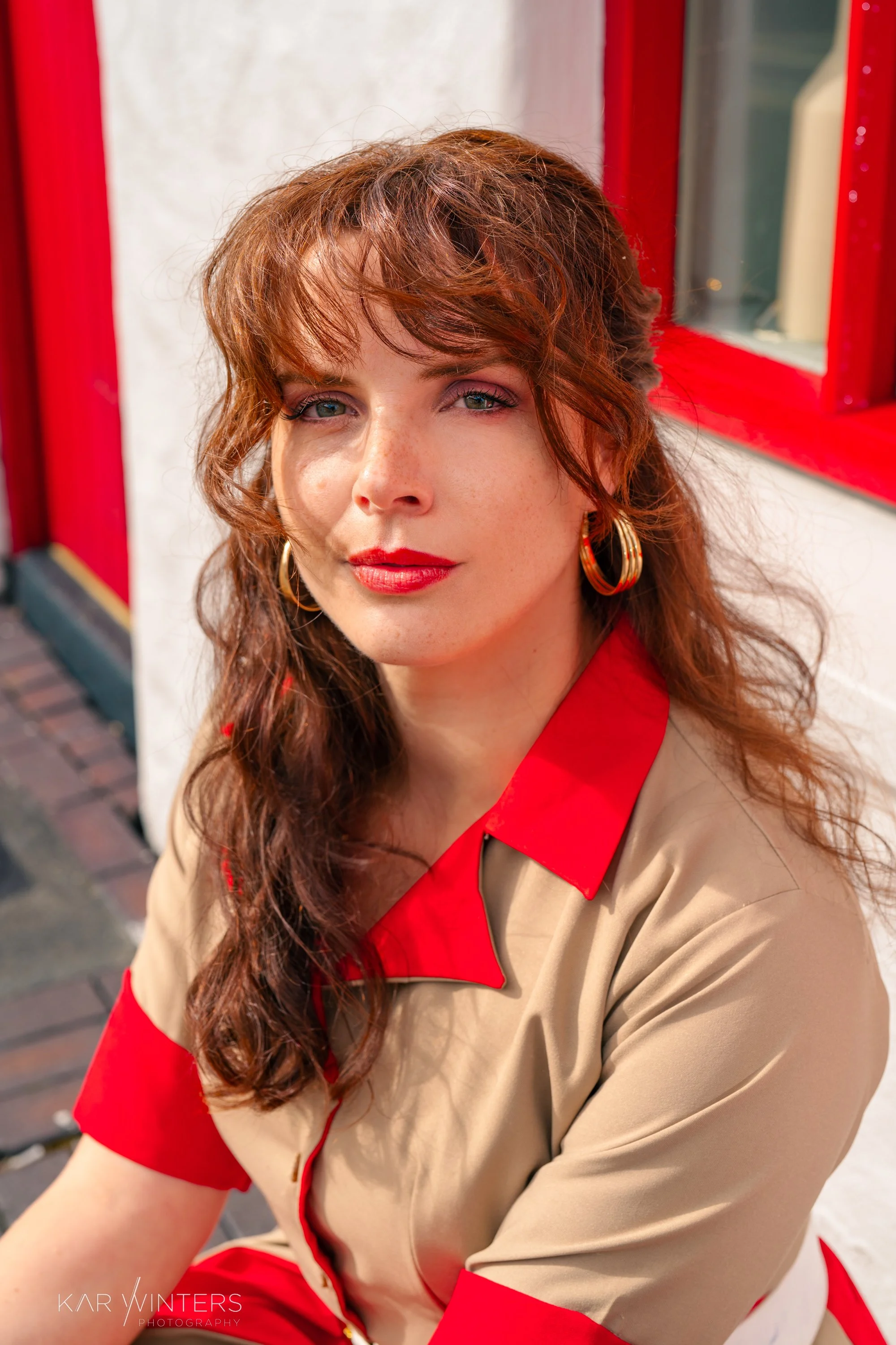 Close-up of a woman with long, curly auburn hair and blue eyes, wearing red lipstick, red earrings, and a beige shirt with red accents, sitting near a red and white building exterior.