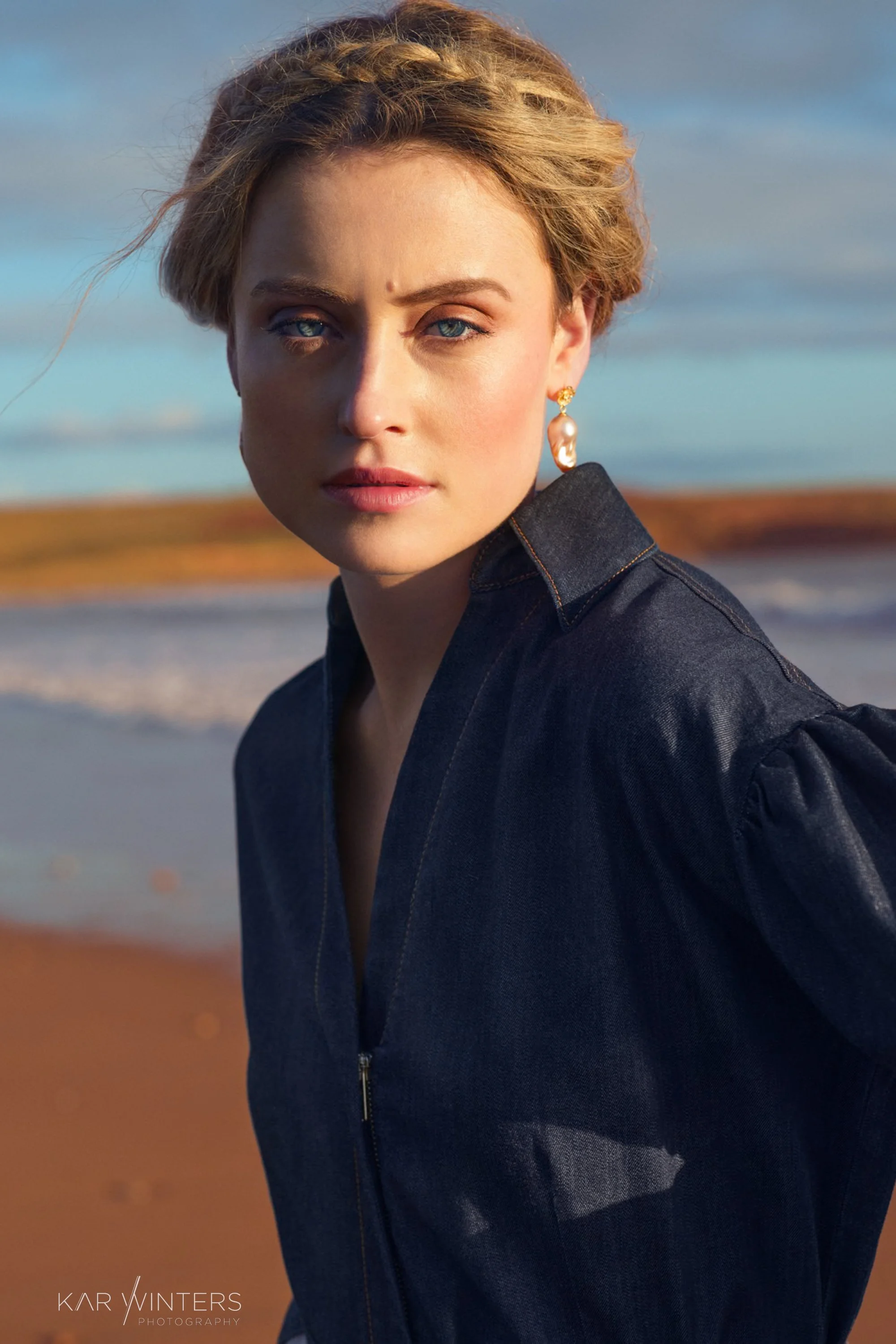 A woman with short, wavy blonde hair and blue eyes stands outdoors at a beach with sand and water in the background. She wears a dark denim jacket and gold earrings with a large pearl. The photo is taken during golden hour, with soft sunlight illumin