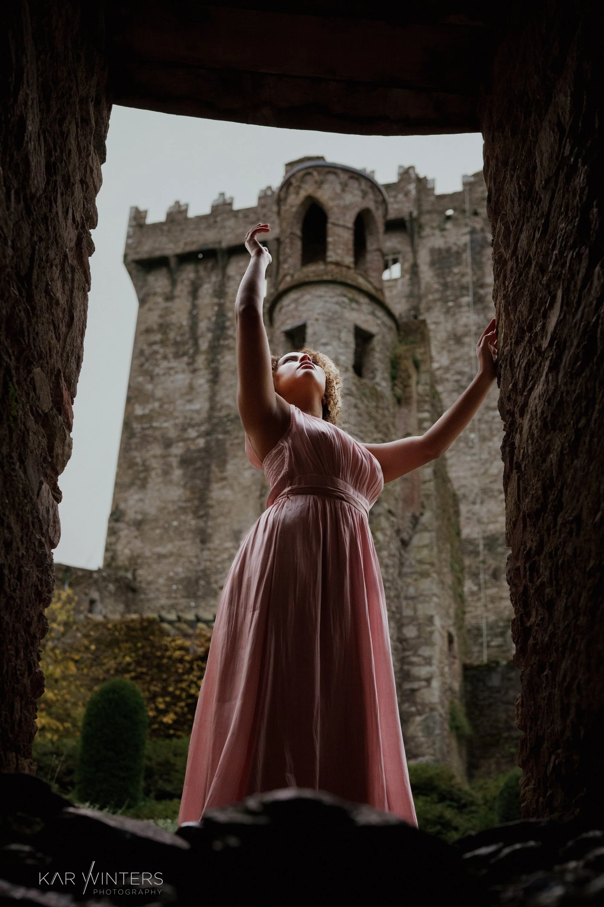A woman in a pink gown standing in a stone archway, looking up and reaching towards the sky, with an old castle in the background.
